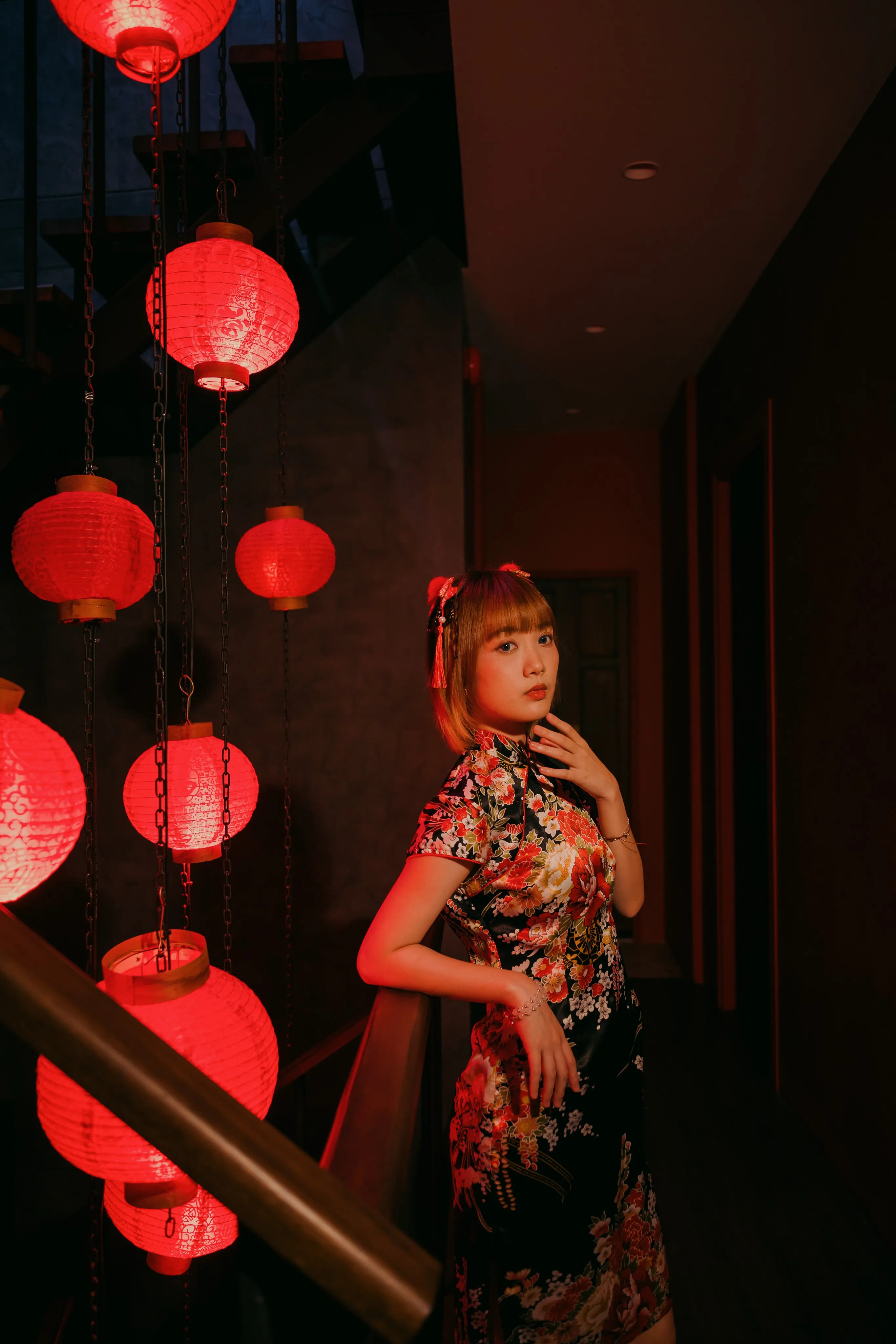 A woman in a black floral qipao posing on a dark staircase hung with red paper lanterns
