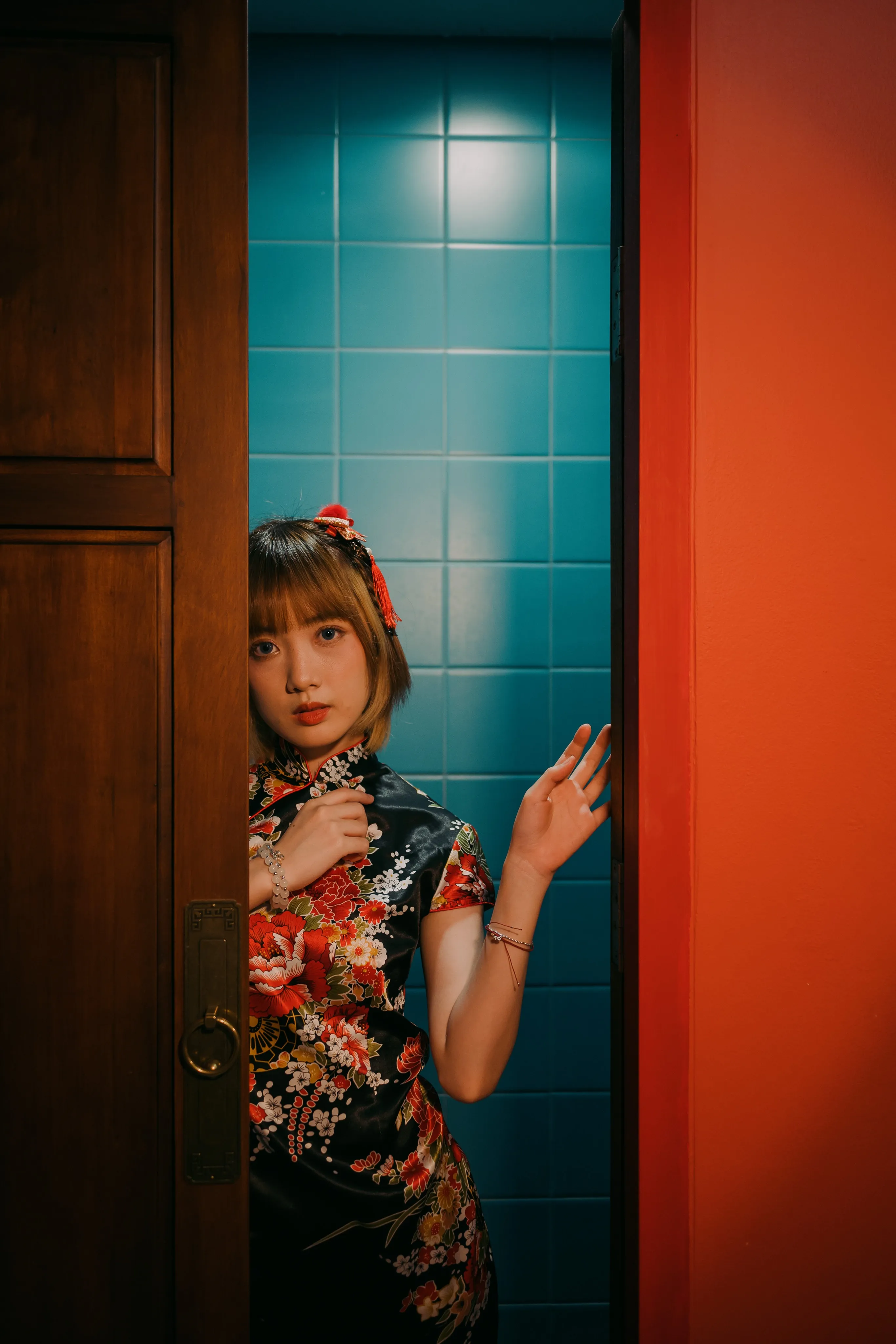 A woman in a black floral qipao peeking out from a wooden doorway against blue tiles and a red wall