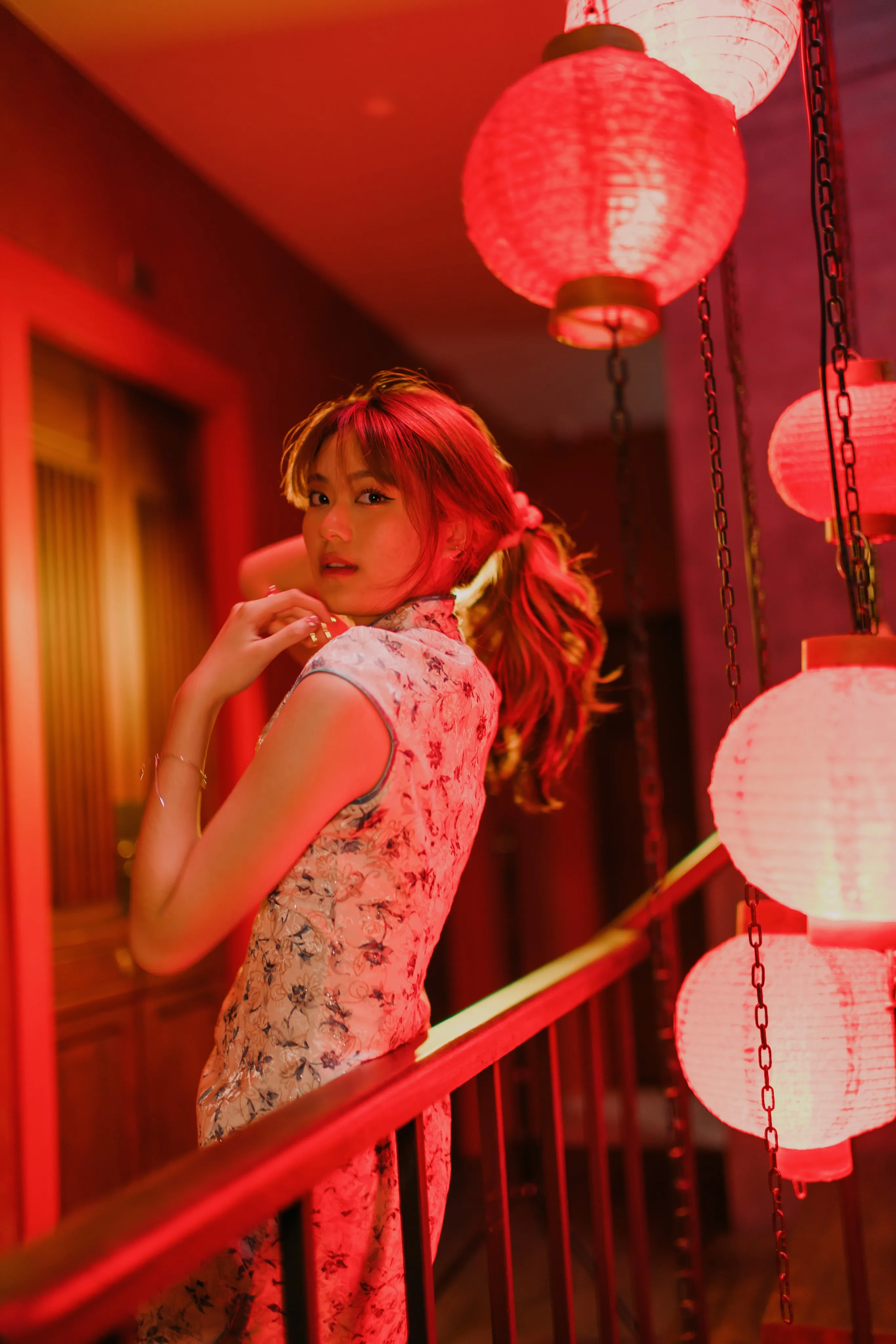 A woman in a floral qipao looking back over her shoulder beside a railing of glowing red lanterns