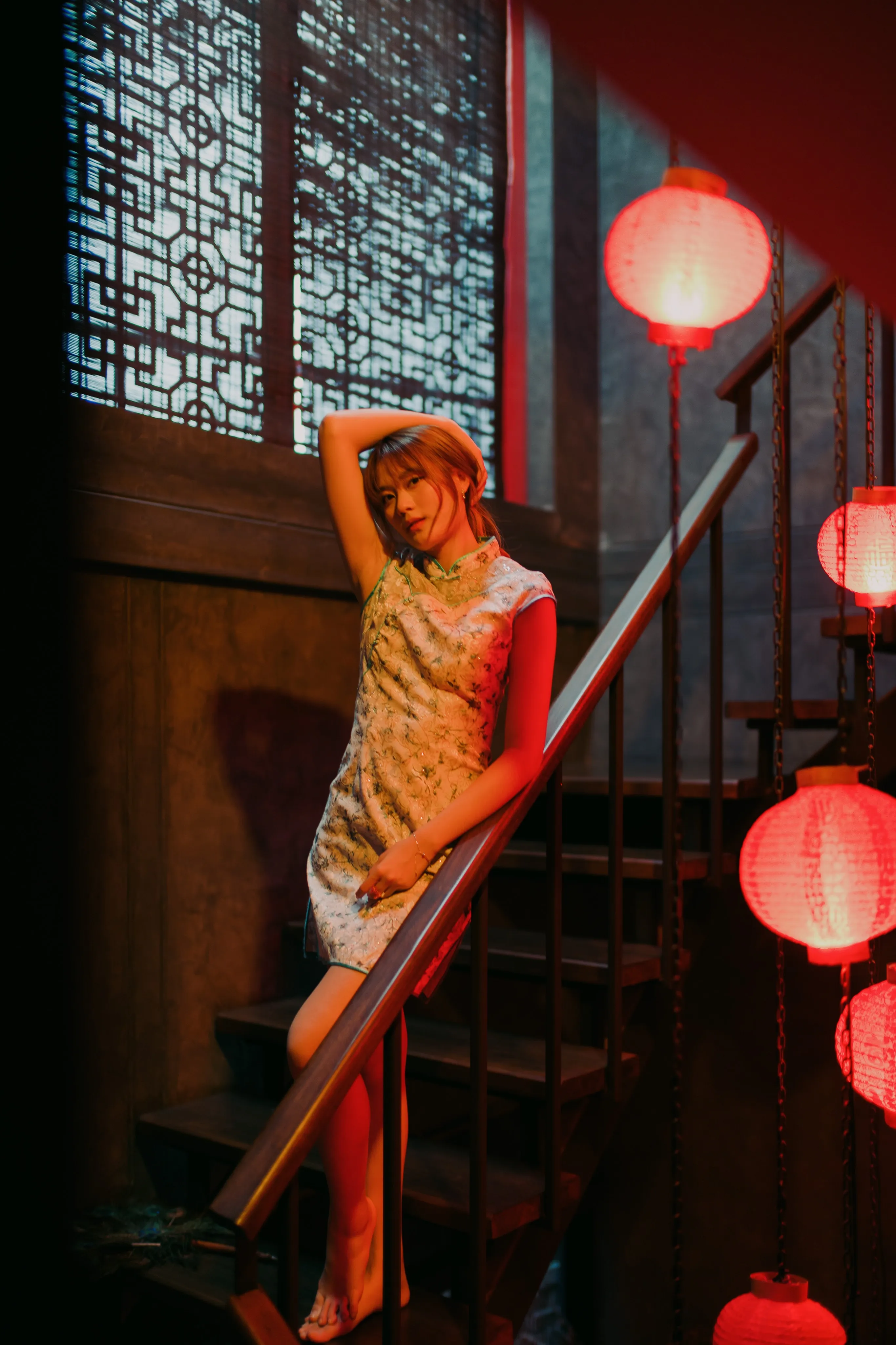 A woman in a floral qipao leaning on a stair railing under a latticed window and red lanterns