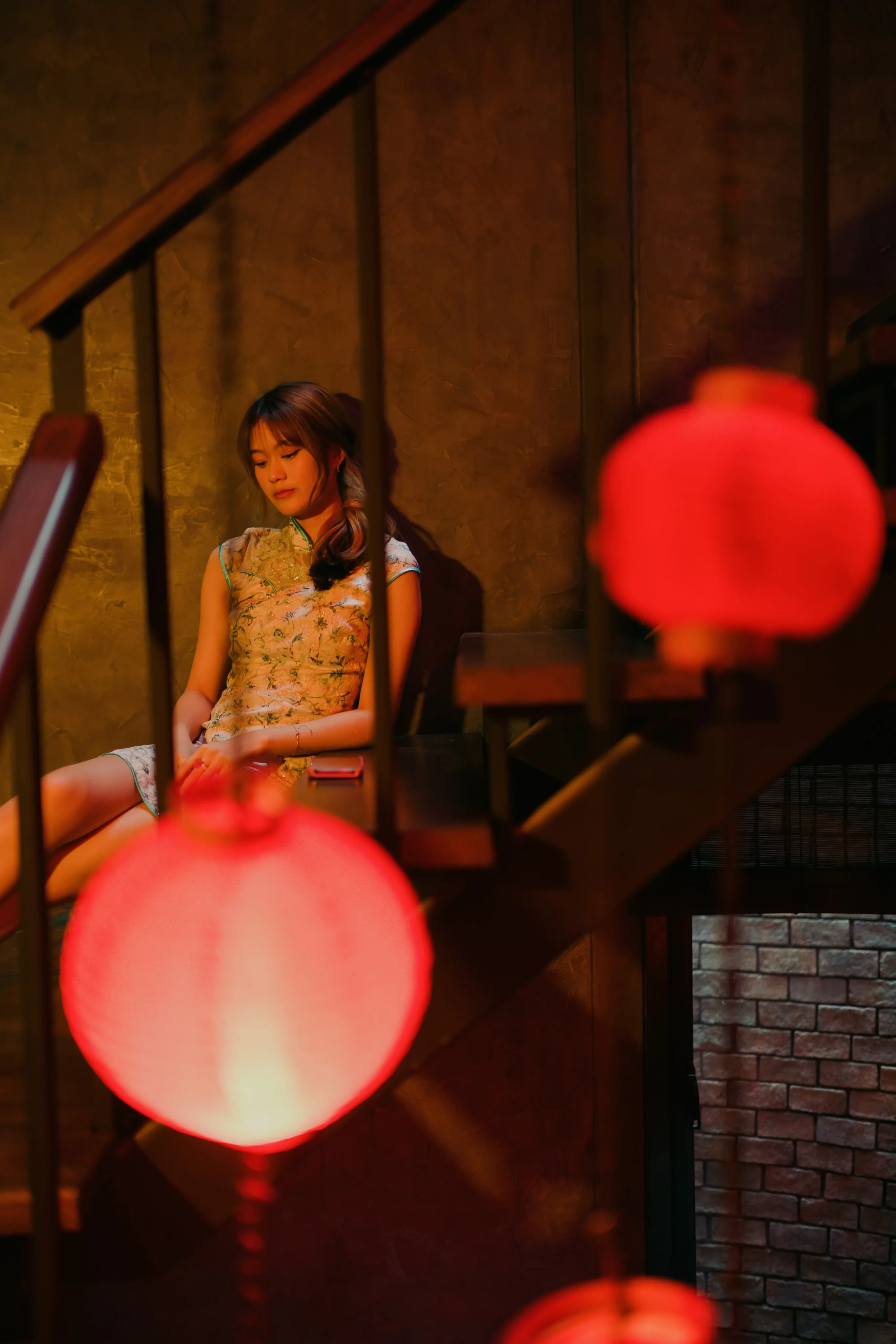 A woman in a qipao seated on a staircase, framed by blurred red lanterns in the foreground