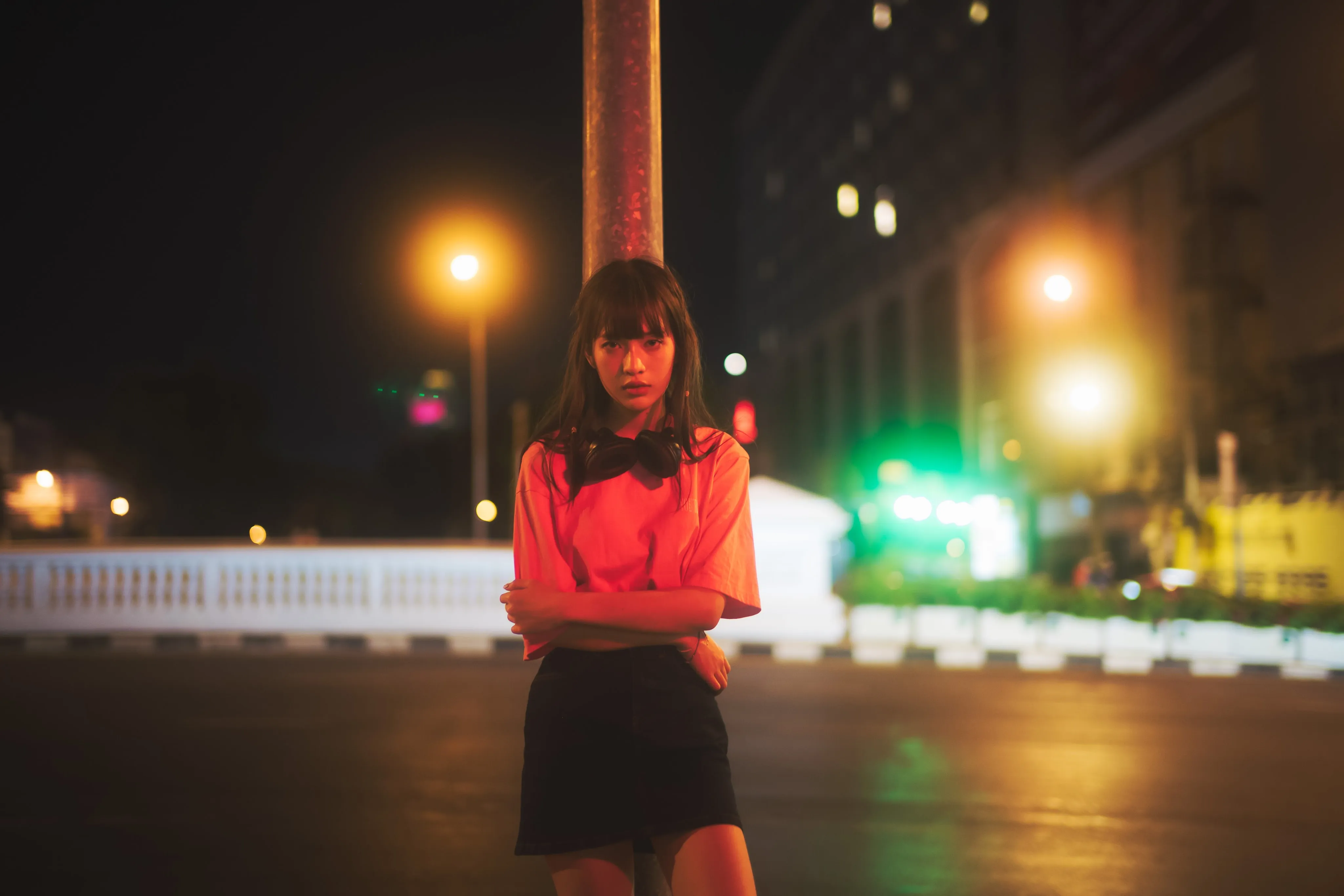 A young woman in a pink T-shirt and black skirt standing with folded arms on a city street at night