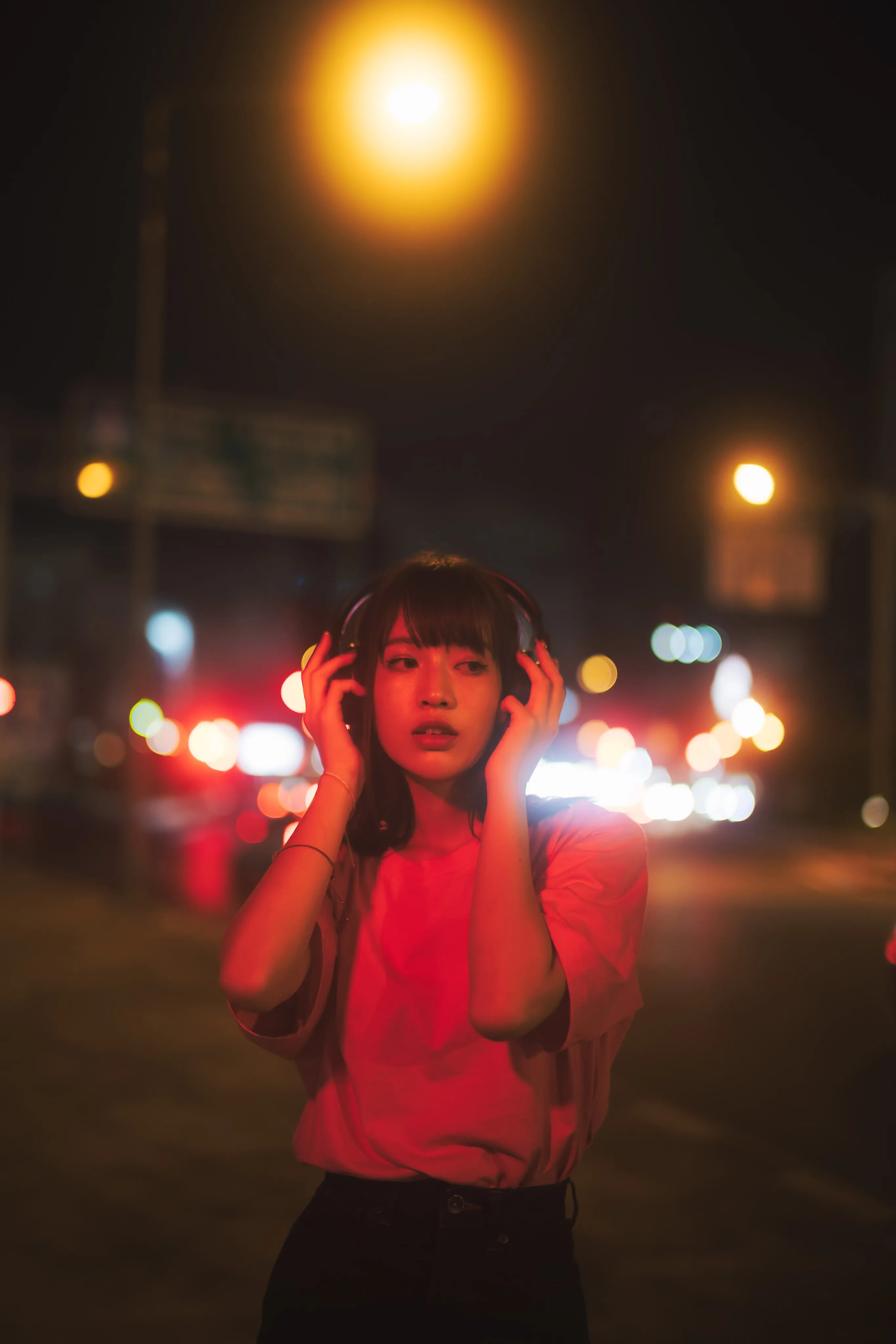 A young woman in a pink T-shirt holding headphones to her ears on a street at night