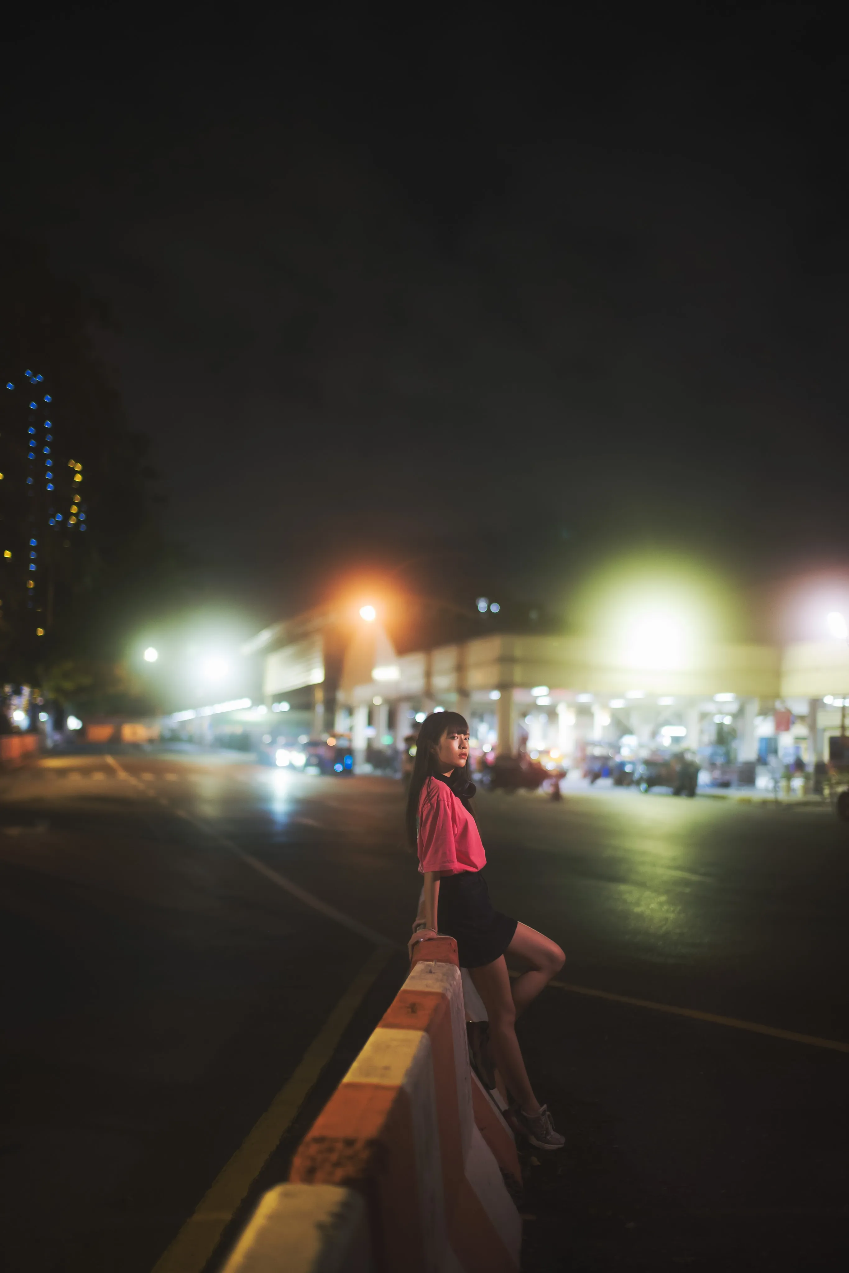 A young woman in a pink T-shirt sitting on a roadside barrier on a wide road at night