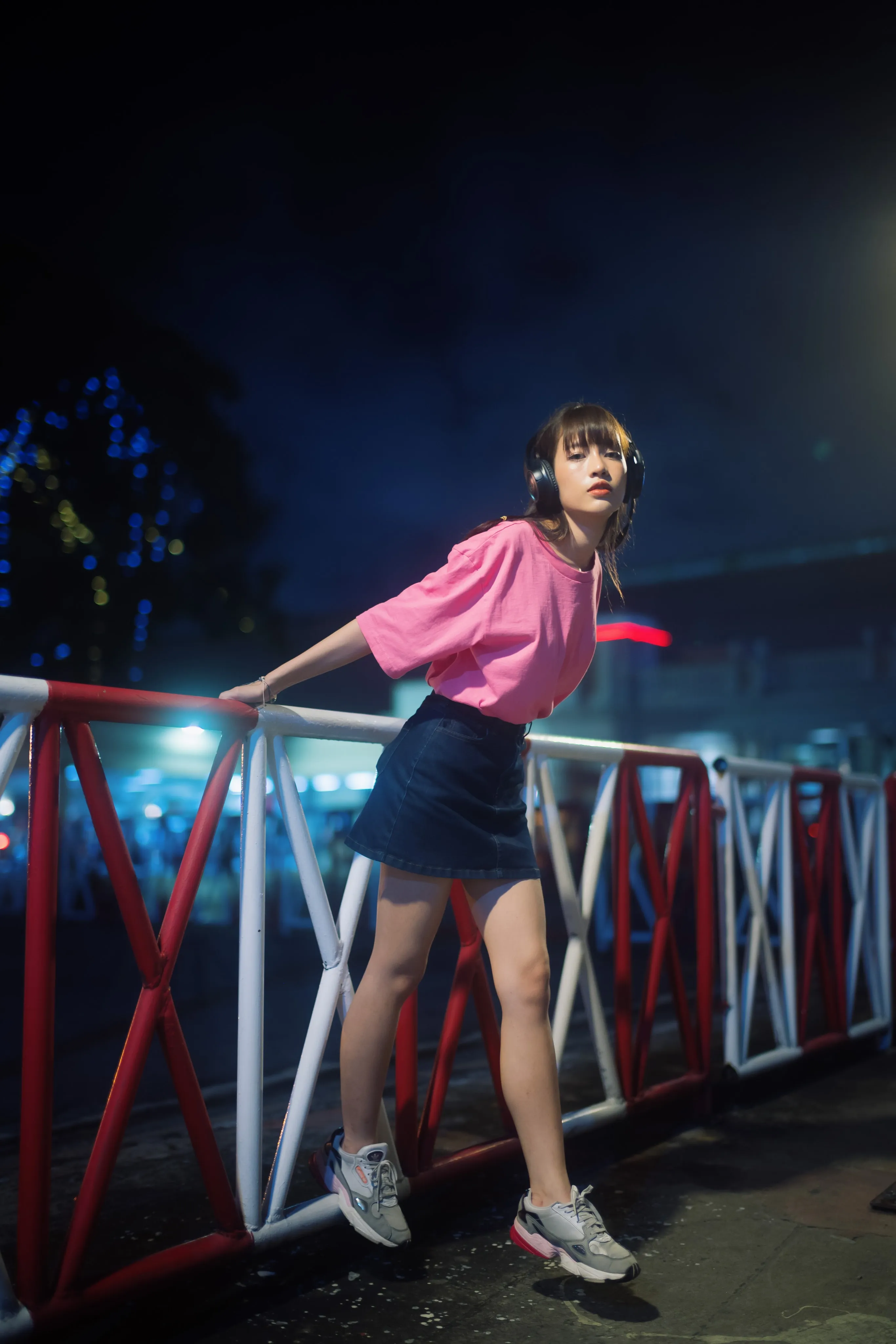 A young woman in a pink T-shirt, denim skirt, and headphones leaning on a red-and-white railing at night