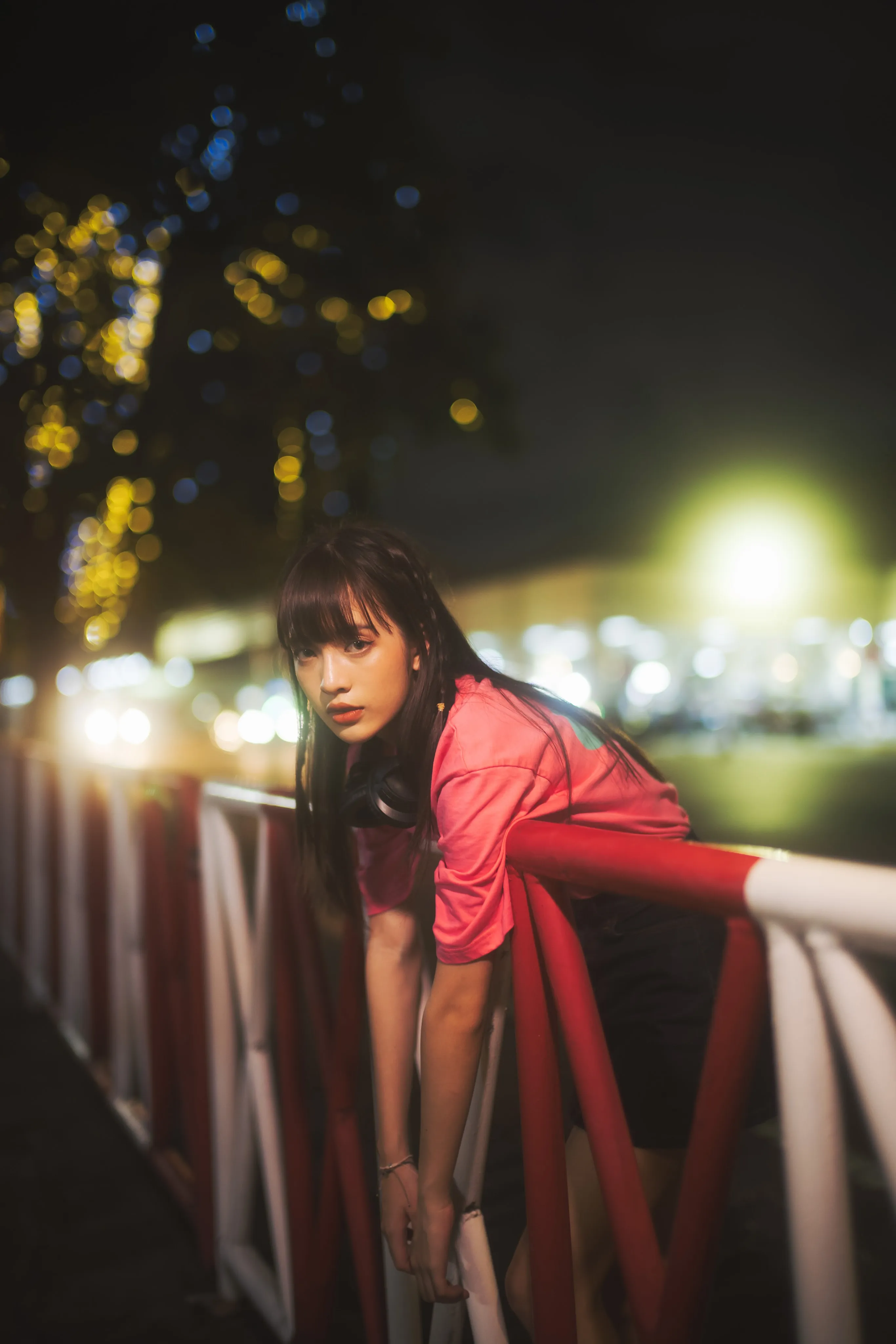 A young woman in a pink T-shirt leaning over a red-and-white railing against blurred night lights