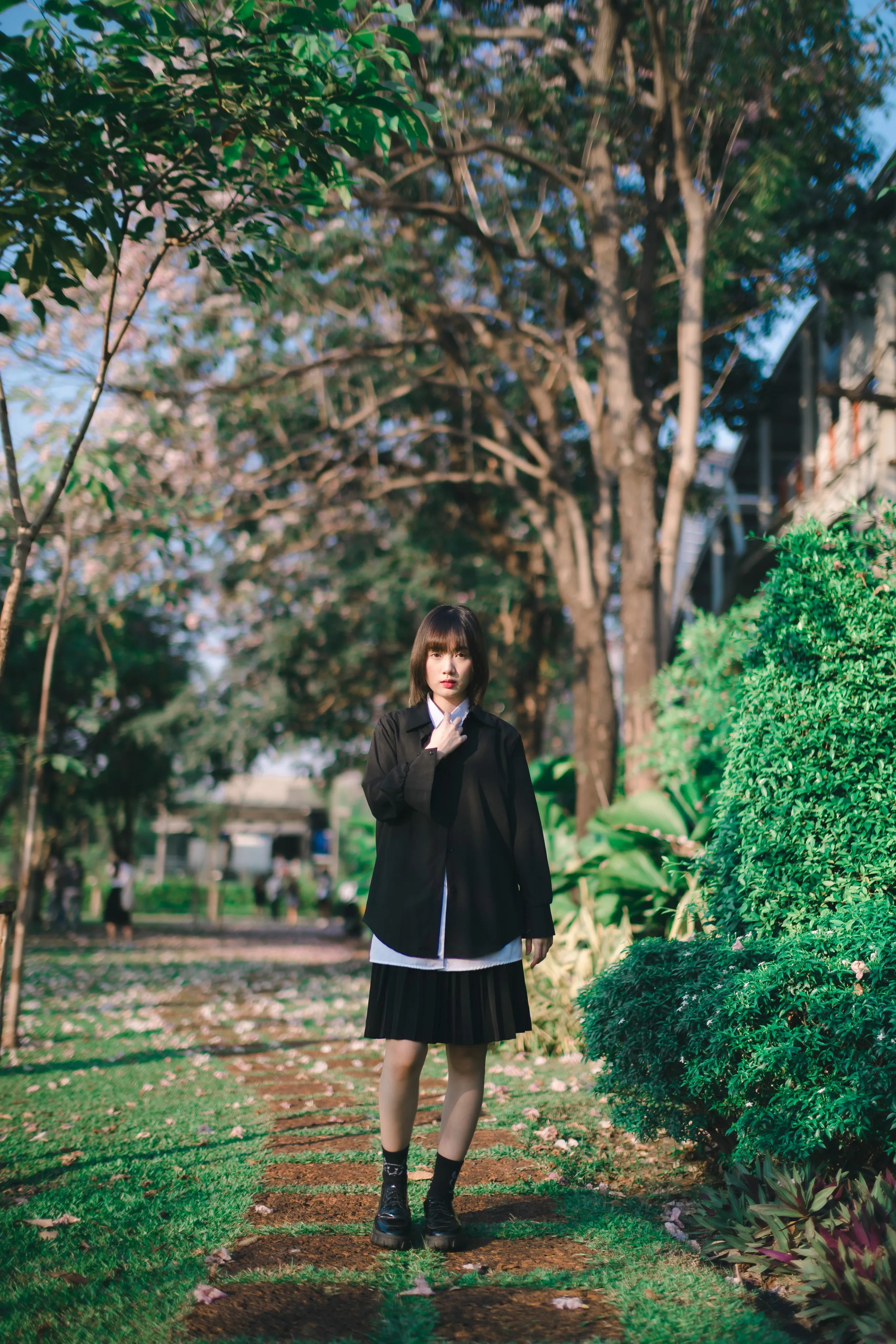 A young woman in a black blazer and pleated skirt standing on a garden path under trees