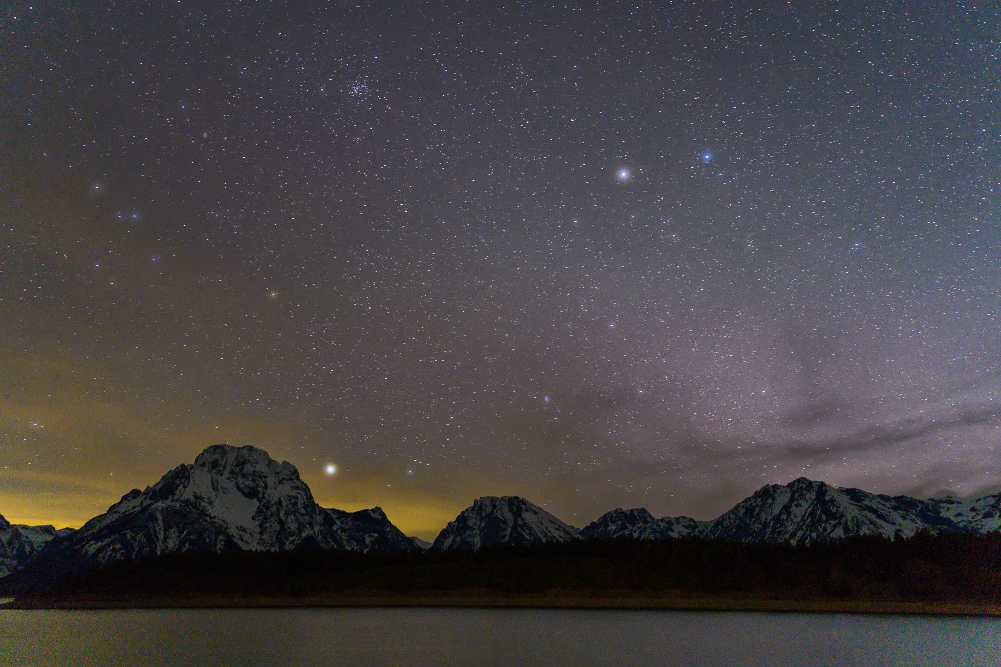 Stars above snow-capped Teton peaks, a dark forested shoreline, and a still lake