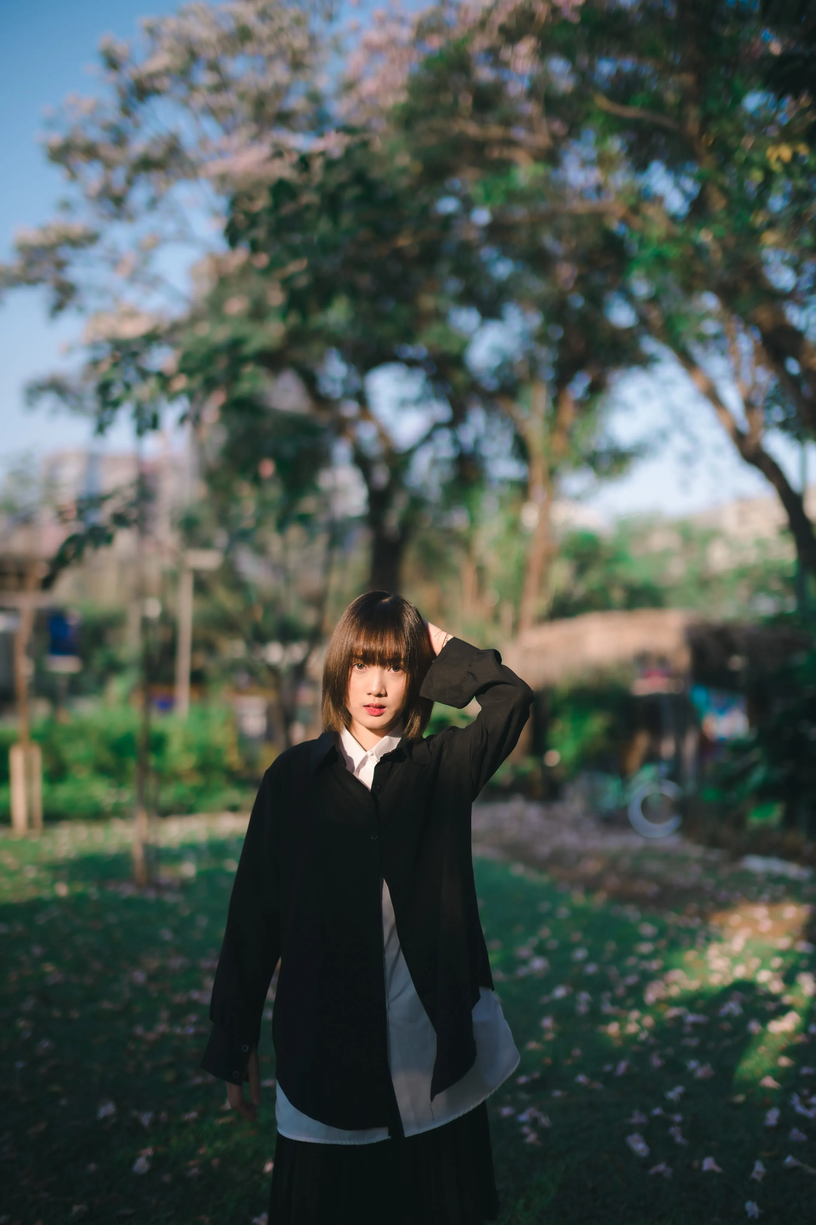 A young woman in an oversized black shirt with a hand in her hair in a sunlit park