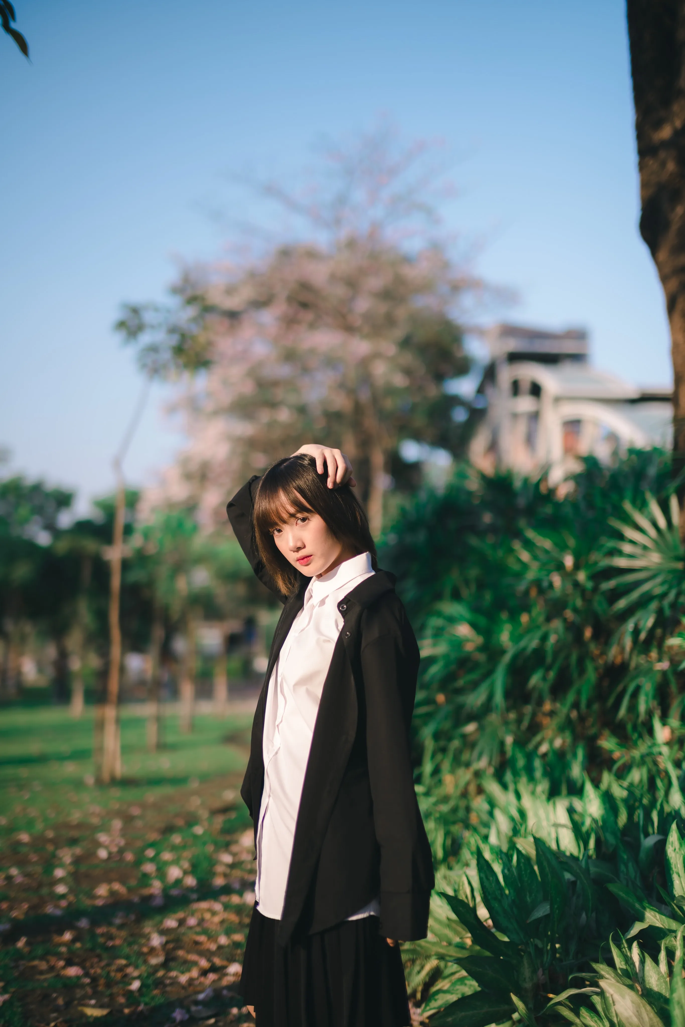 A young woman in a black blazer and white shirt with a hand on her head in a park with blossom trees