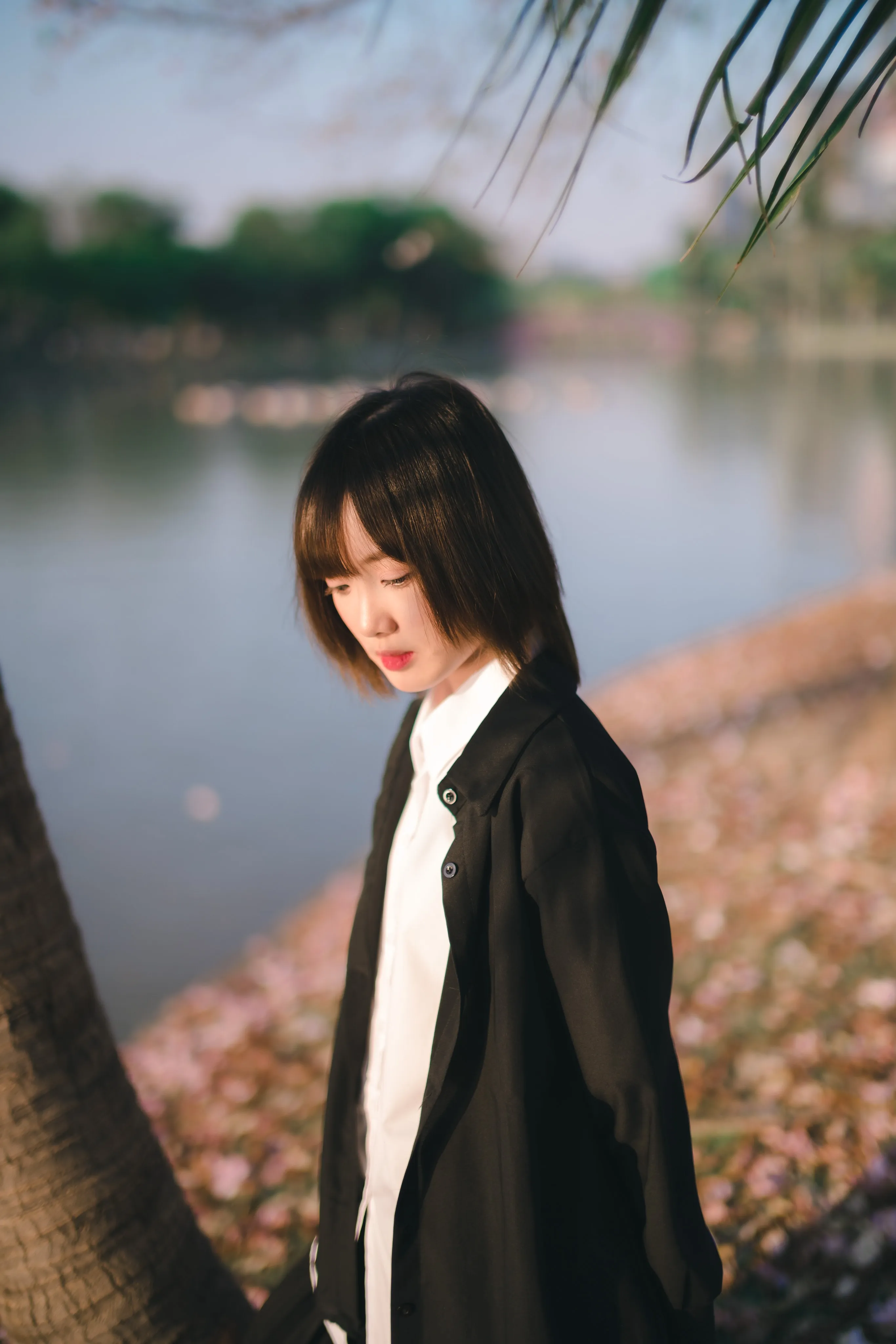 A young woman in a black jacket and white shirt looking down beside a lake edged with fallen pink blossoms