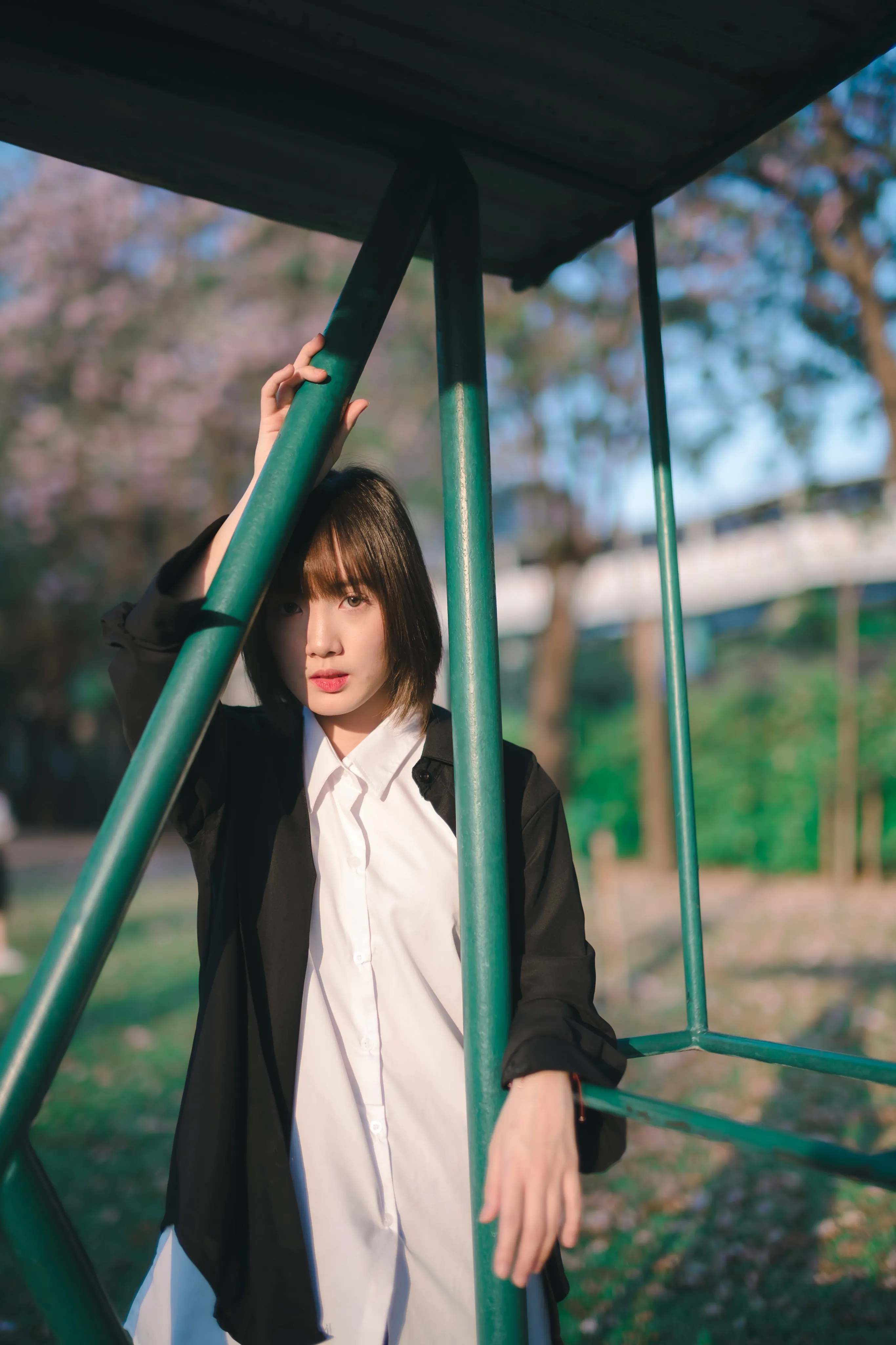 A young woman in a black jacket and white shirt leaning on a green metal frame in a park