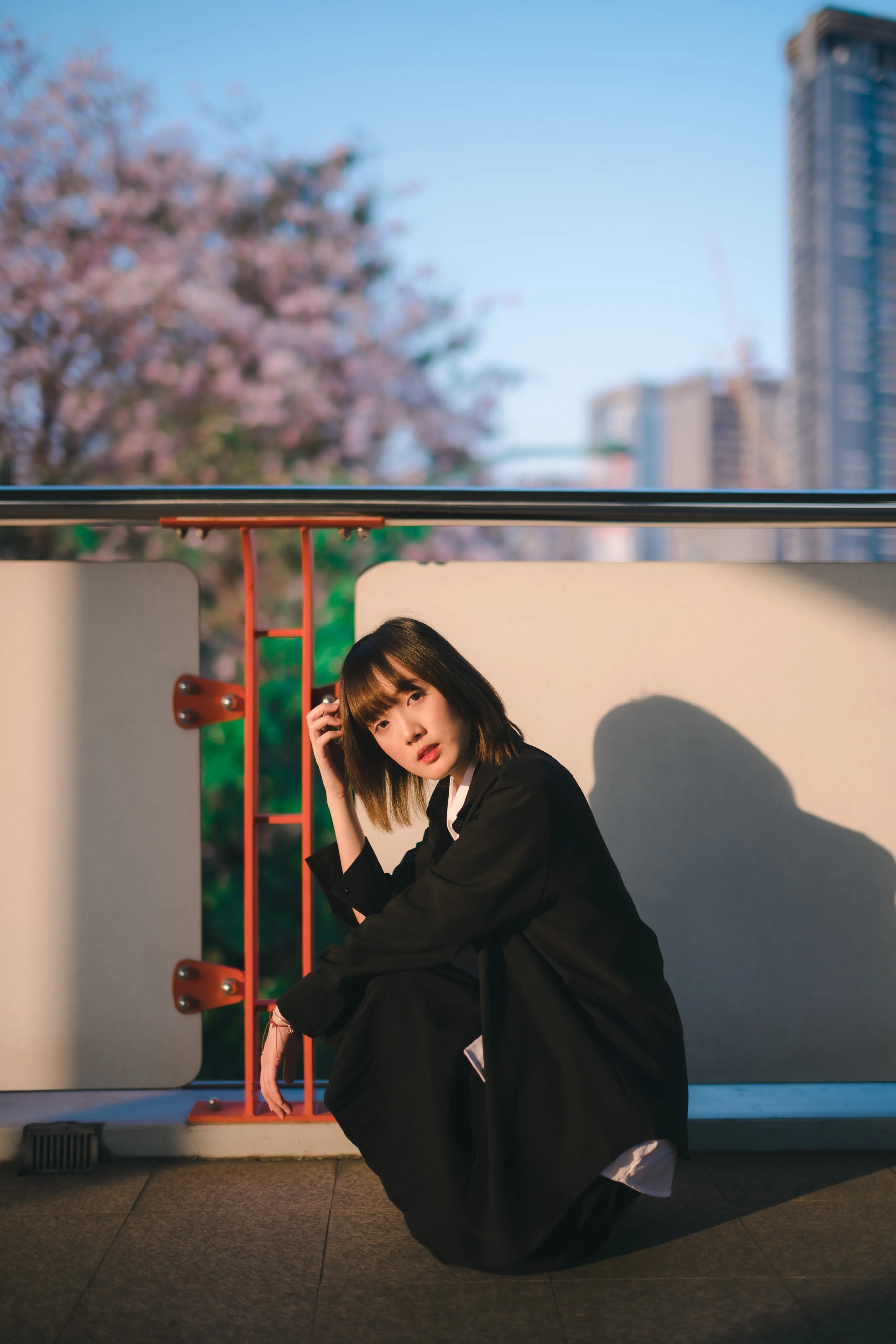 A young woman in a black jacket crouching against a walkway railing in warm evening light
