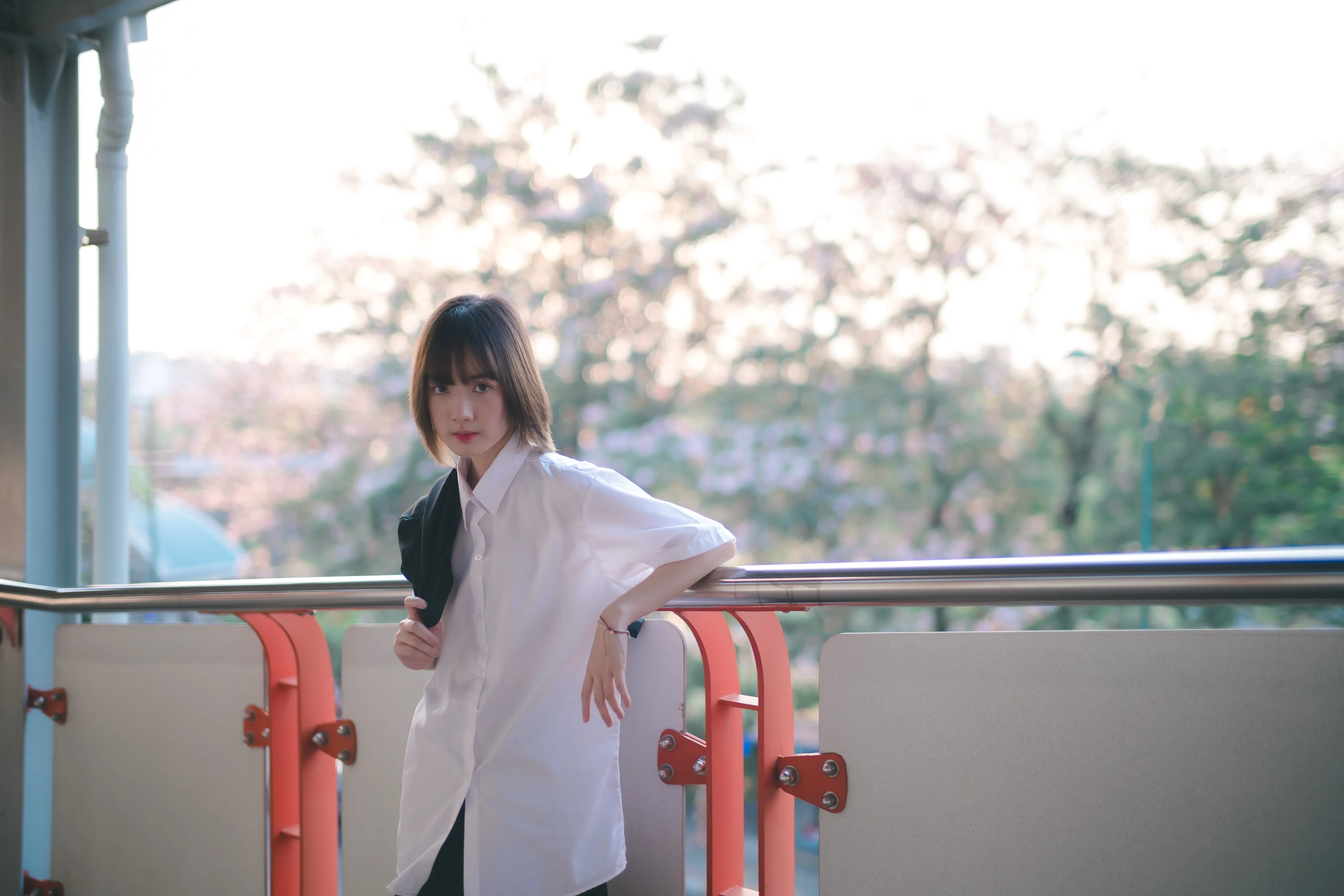 A young woman in a white shirt leaning on a walkway railing with blossom trees behind her