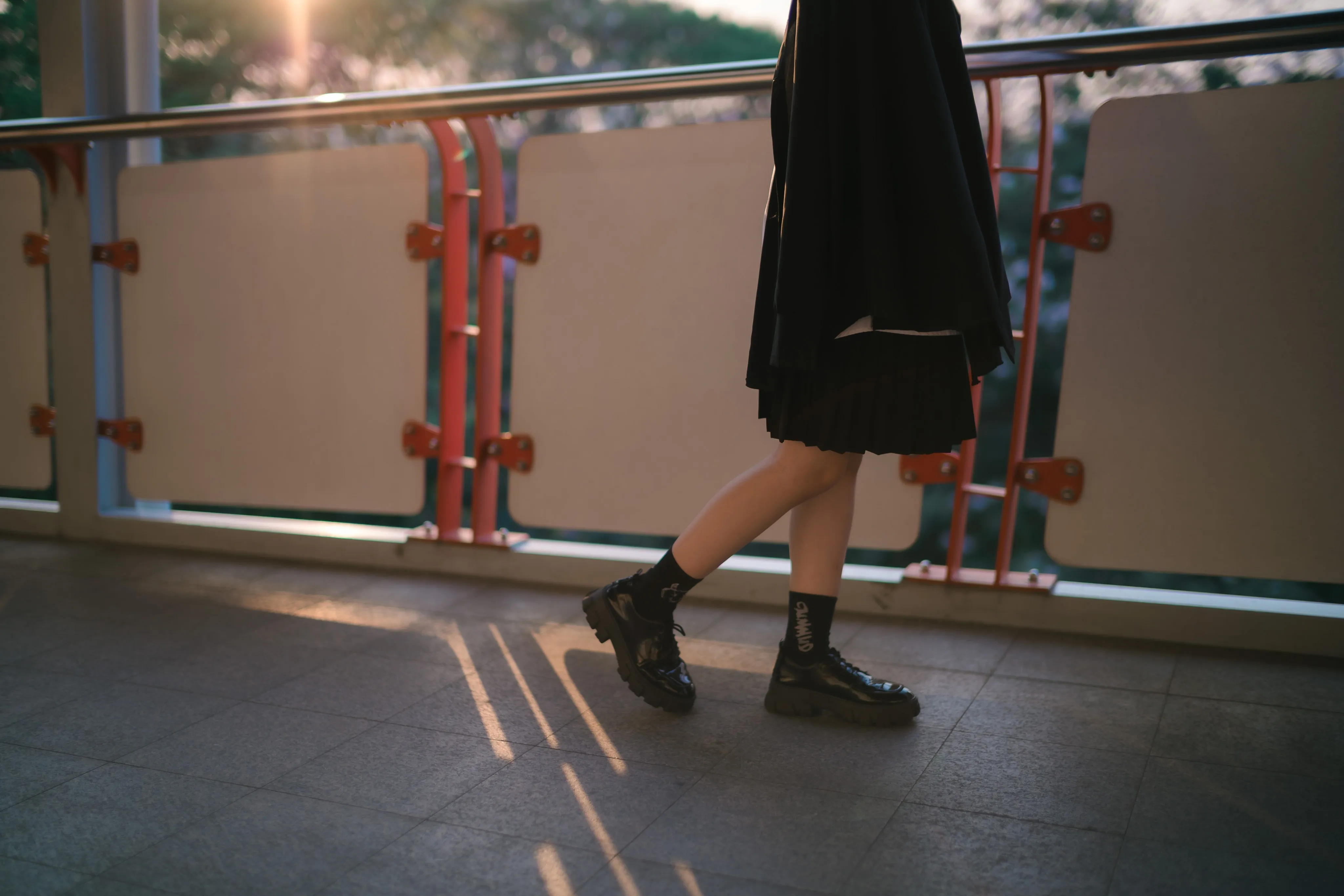 Legs in black boots and a pleated skirt stepping across a sunlit covered walkway