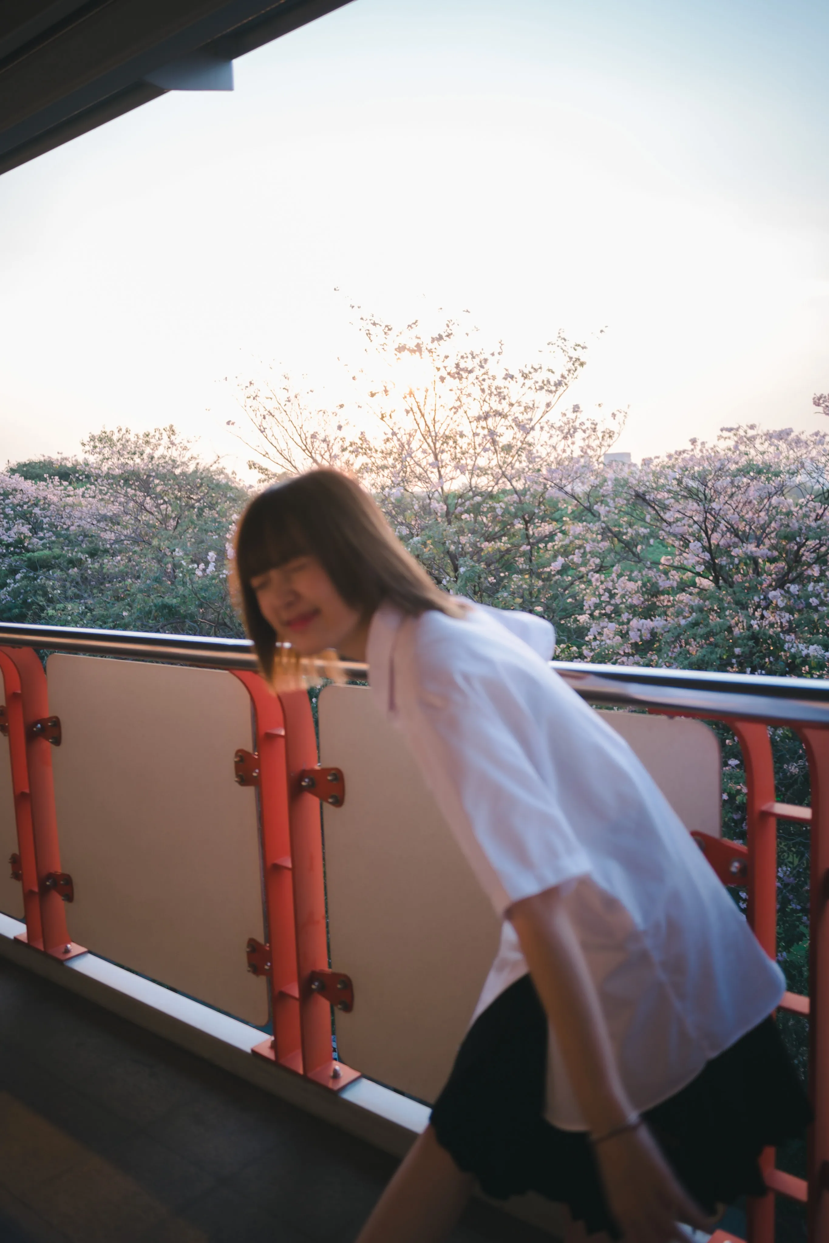A young woman in a white shirt and pleated skirt leaning over a walkway railing, blurred in motion at sunset