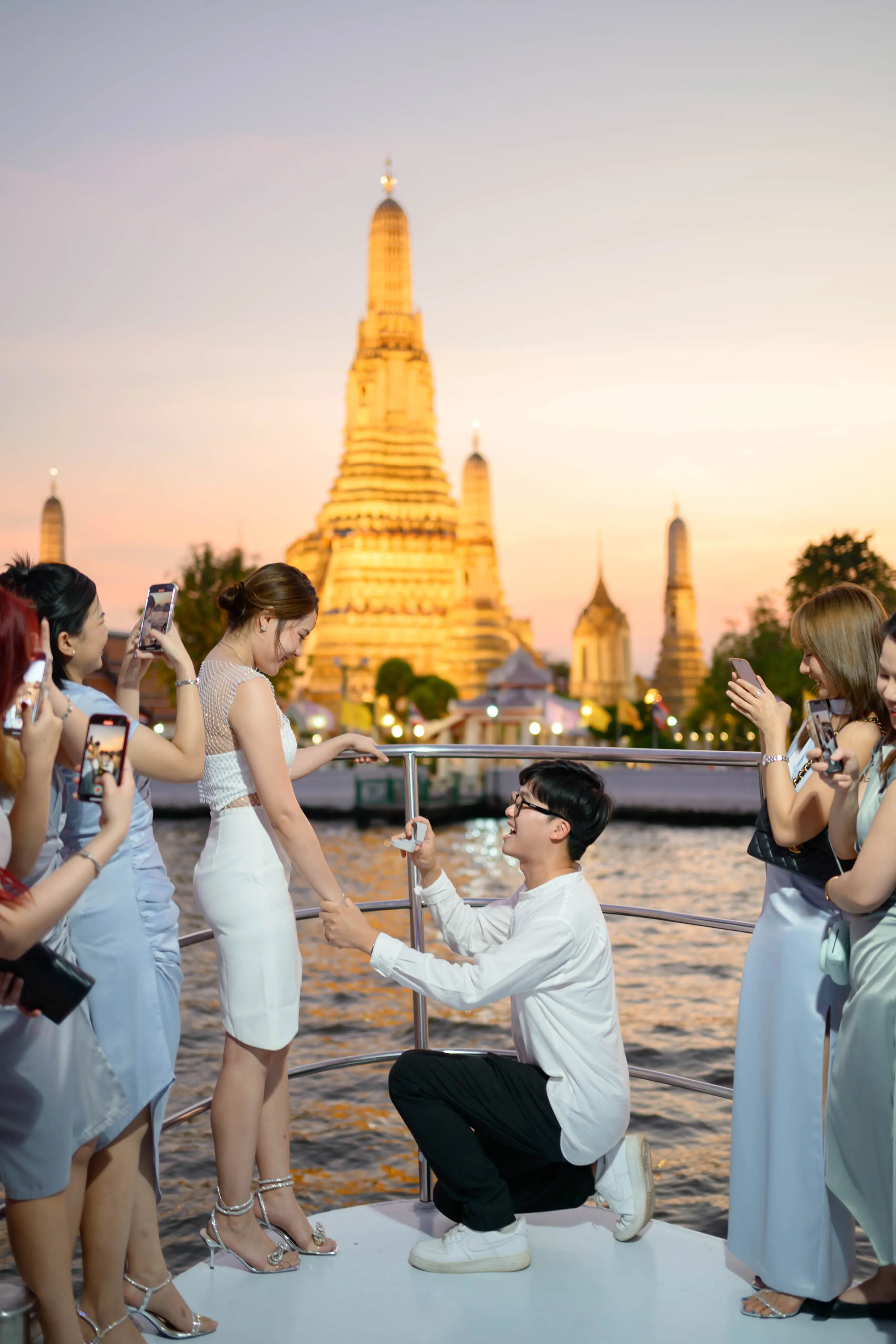A man proposing to a woman in front of a riverside temple at sunset