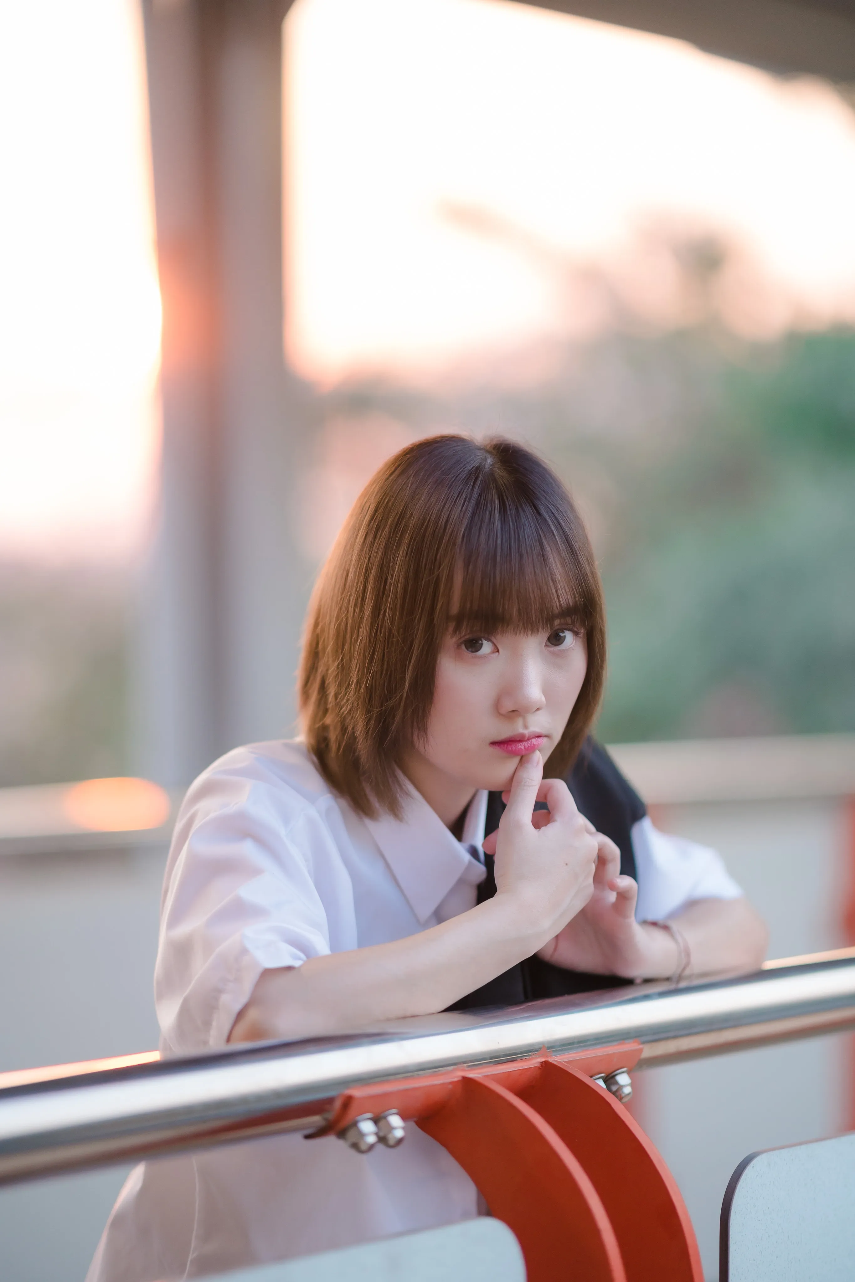 A young woman in a white shirt resting her chin on her hand at a walkway railing at sunset