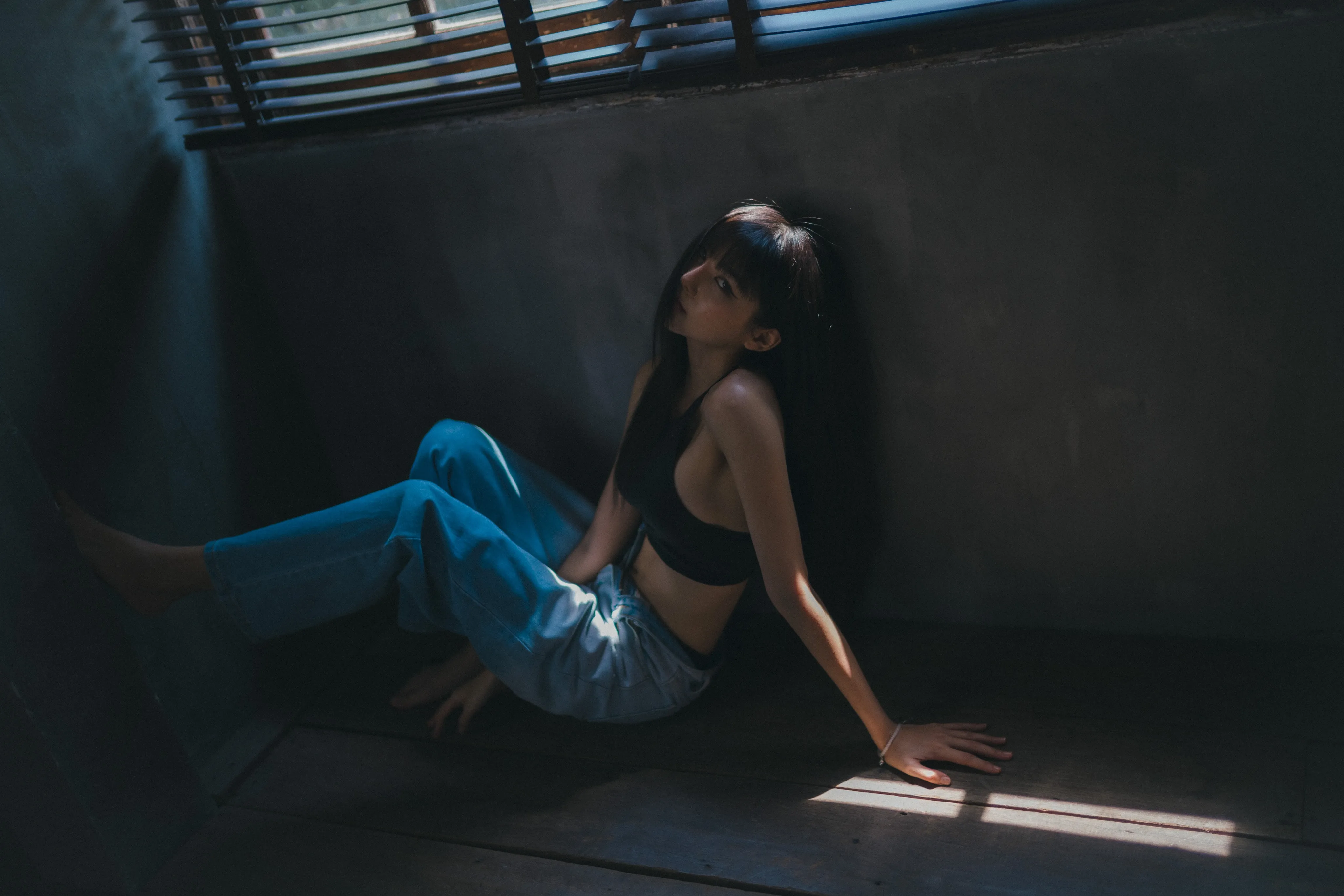 A woman in a black crop top and jeans sitting on the floor in a dim room, looking up toward window blinds