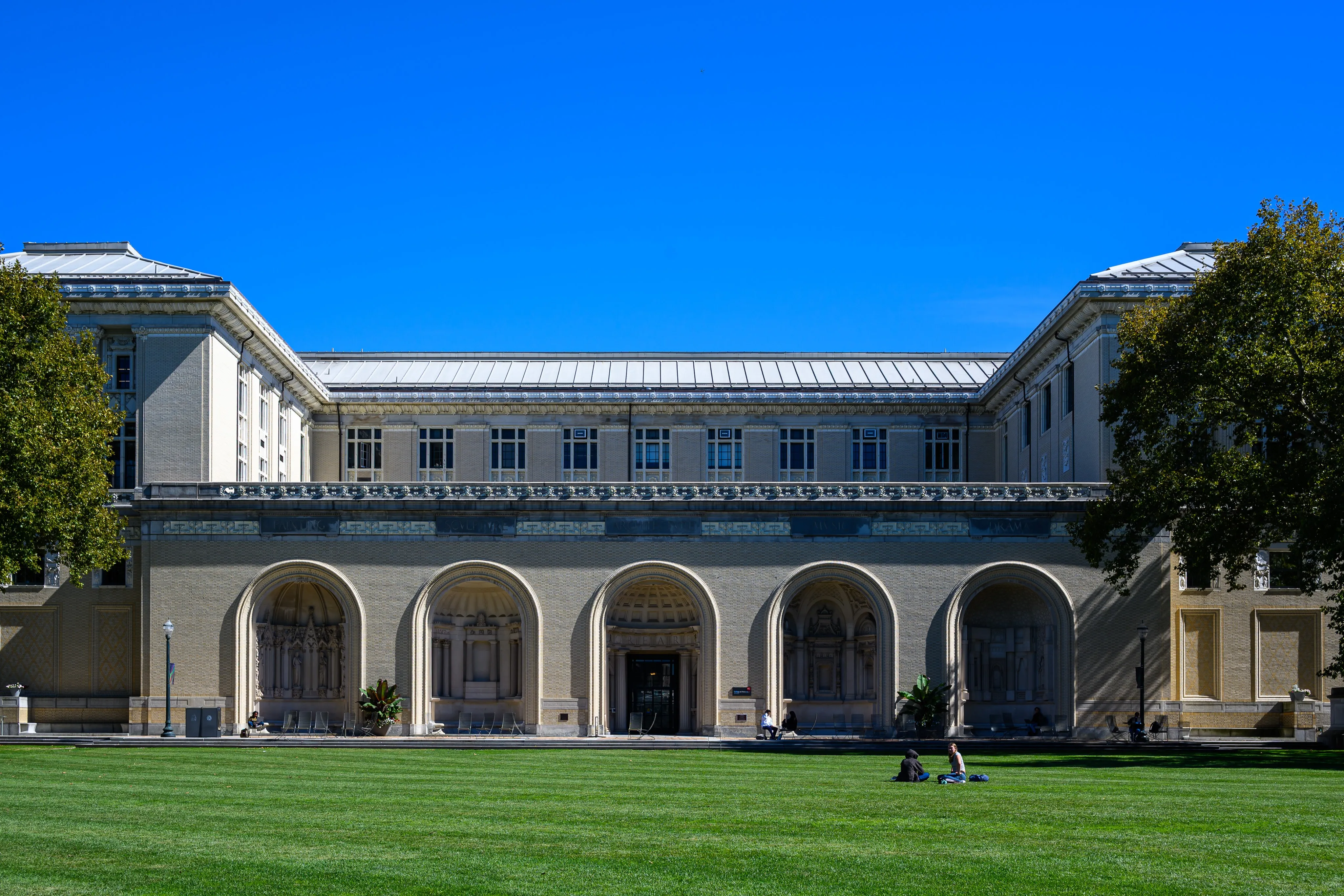 The arched Beaux-Arts facade of a limestone building with five ornate niches, facing a wide green lawn where a few students sit, under a clear blue sky.