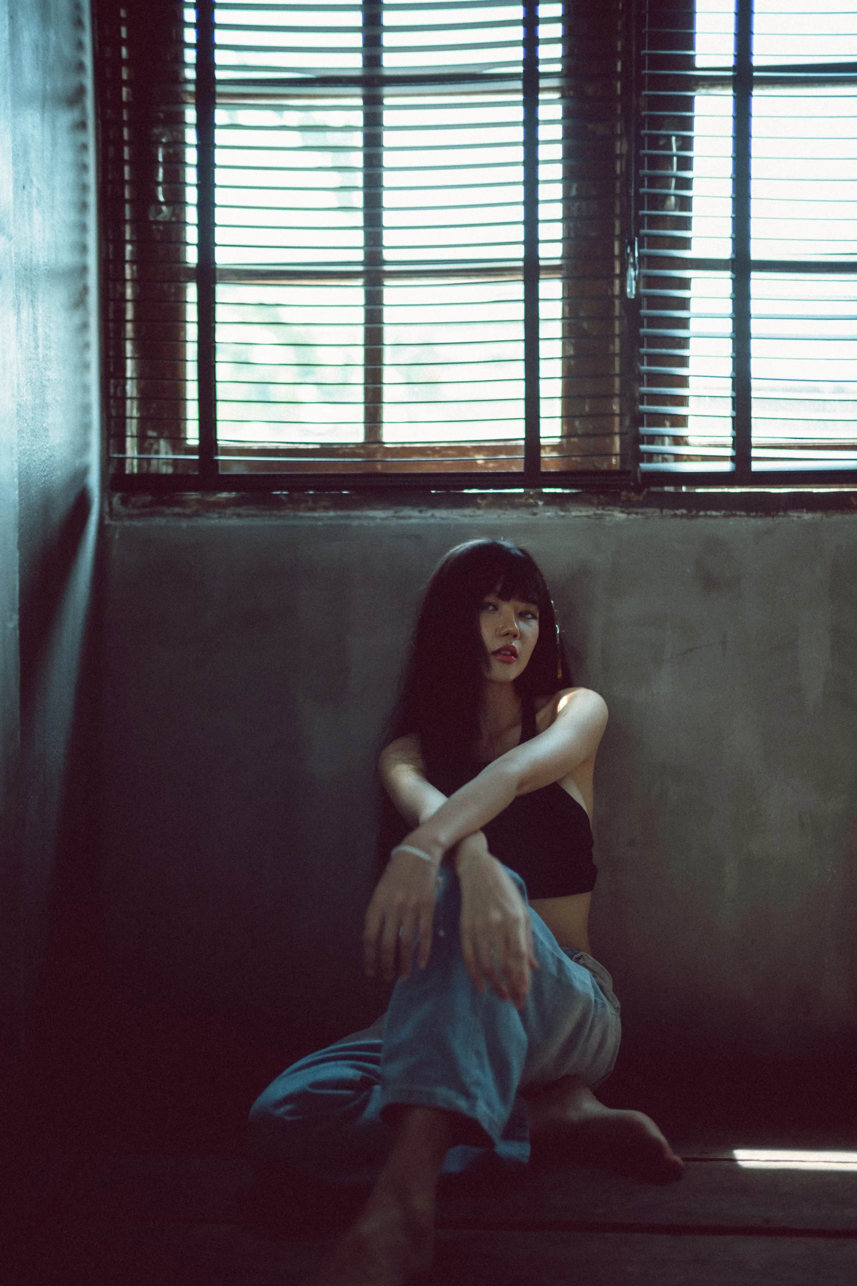 A woman in a black crop top and jeans sitting against a concrete wall beneath a window with blinds