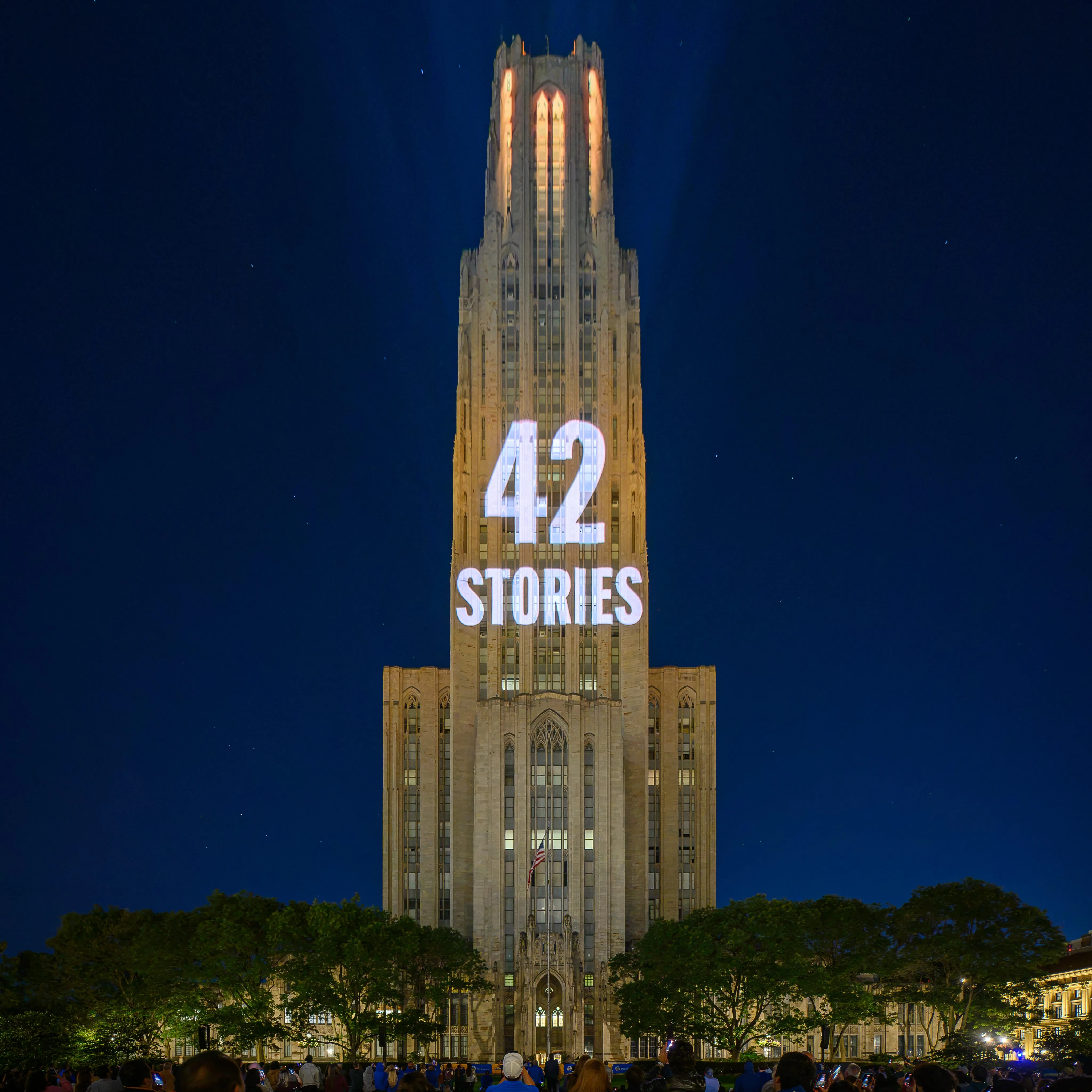 The Cathedral of Learning illuminated at night with “42 STORIES” projected onto its Gothic tower, above trees and a crowd watching the centennial light show.