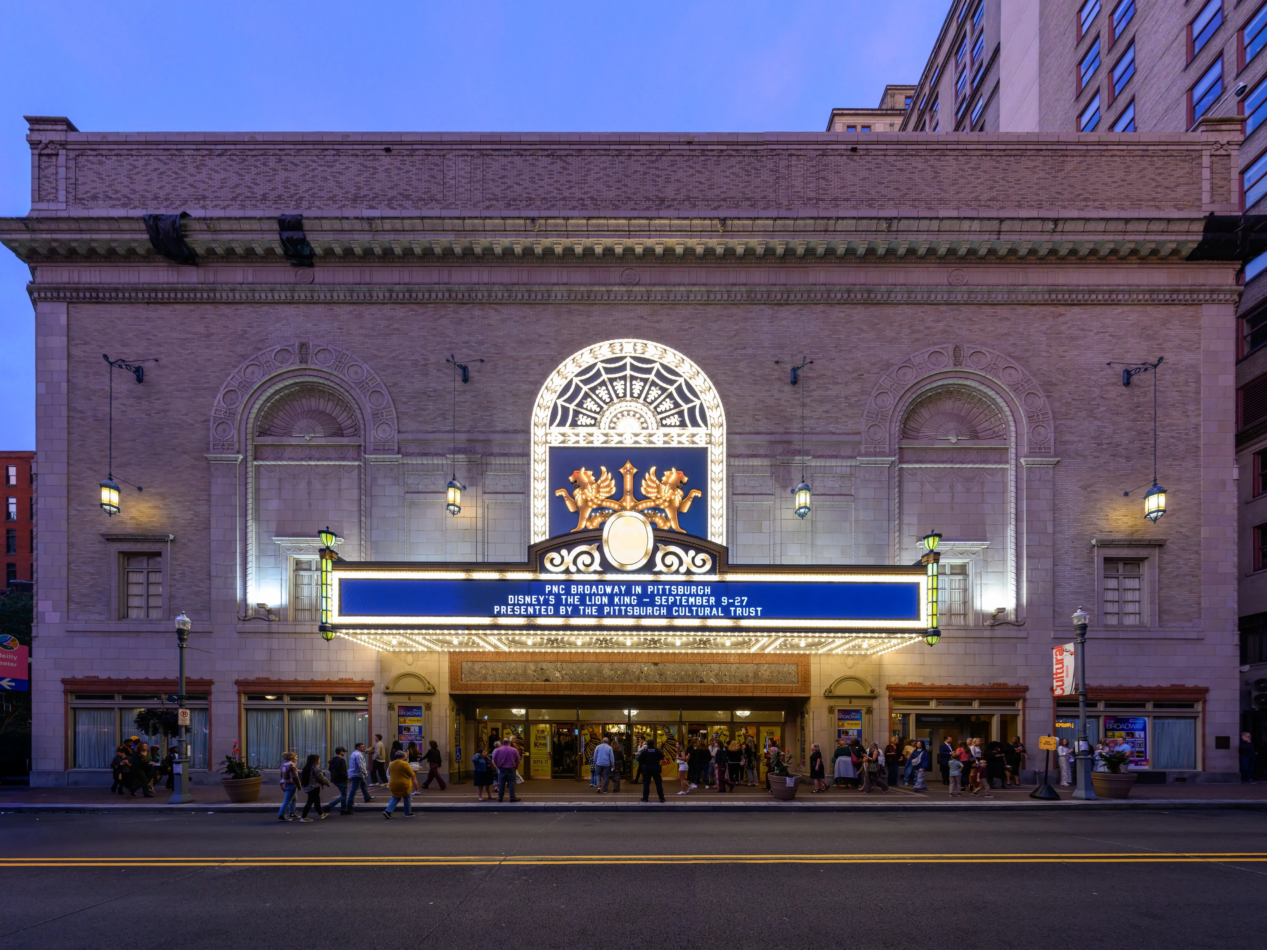 The ornate facade of the Benedum Center at dusk, with an illuminated marquee advertising Disney’s The Lion King and theatergoers gathered outside the entrance.