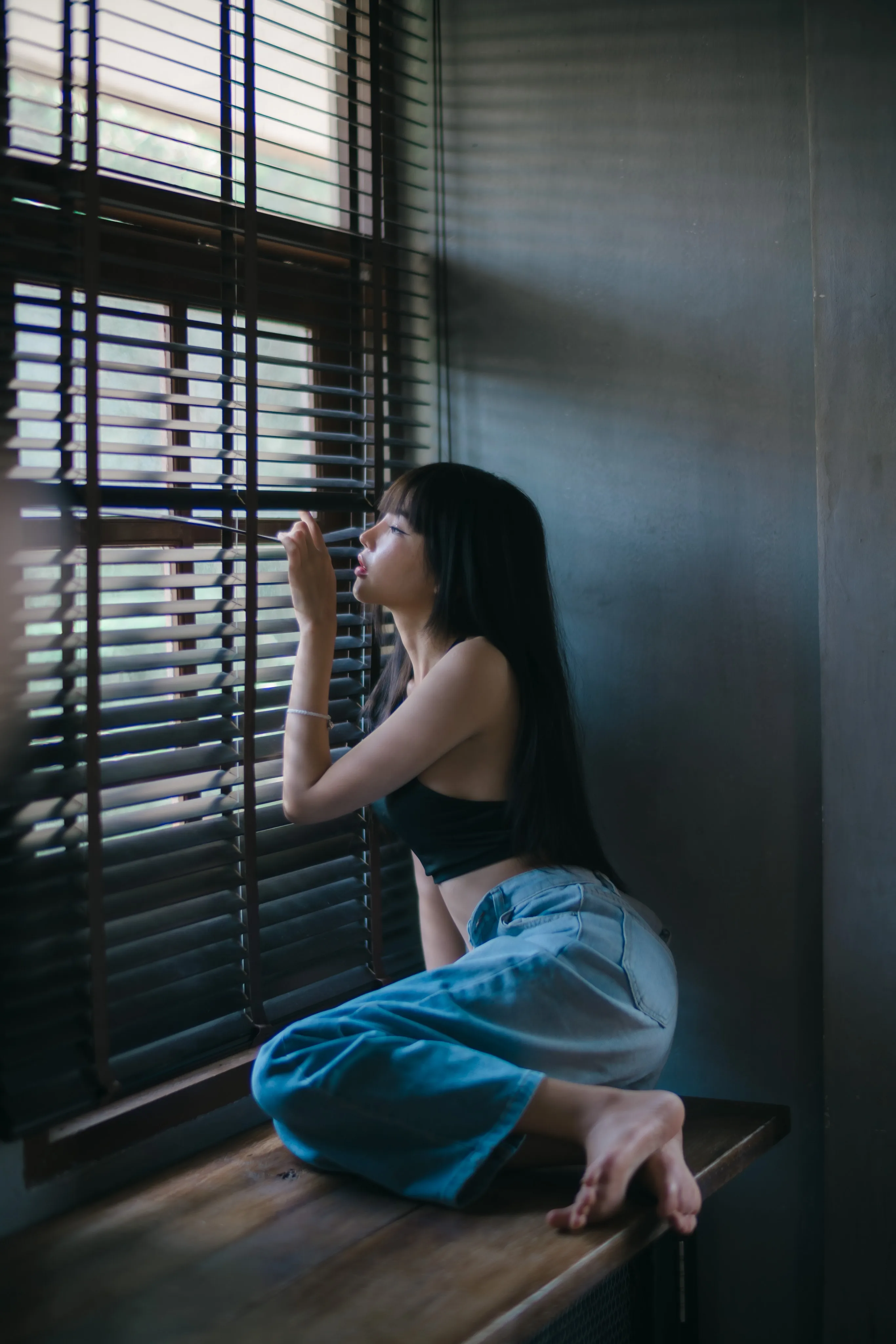 A woman in jeans sitting on a wooden ledge, peering through the slats of a window blind