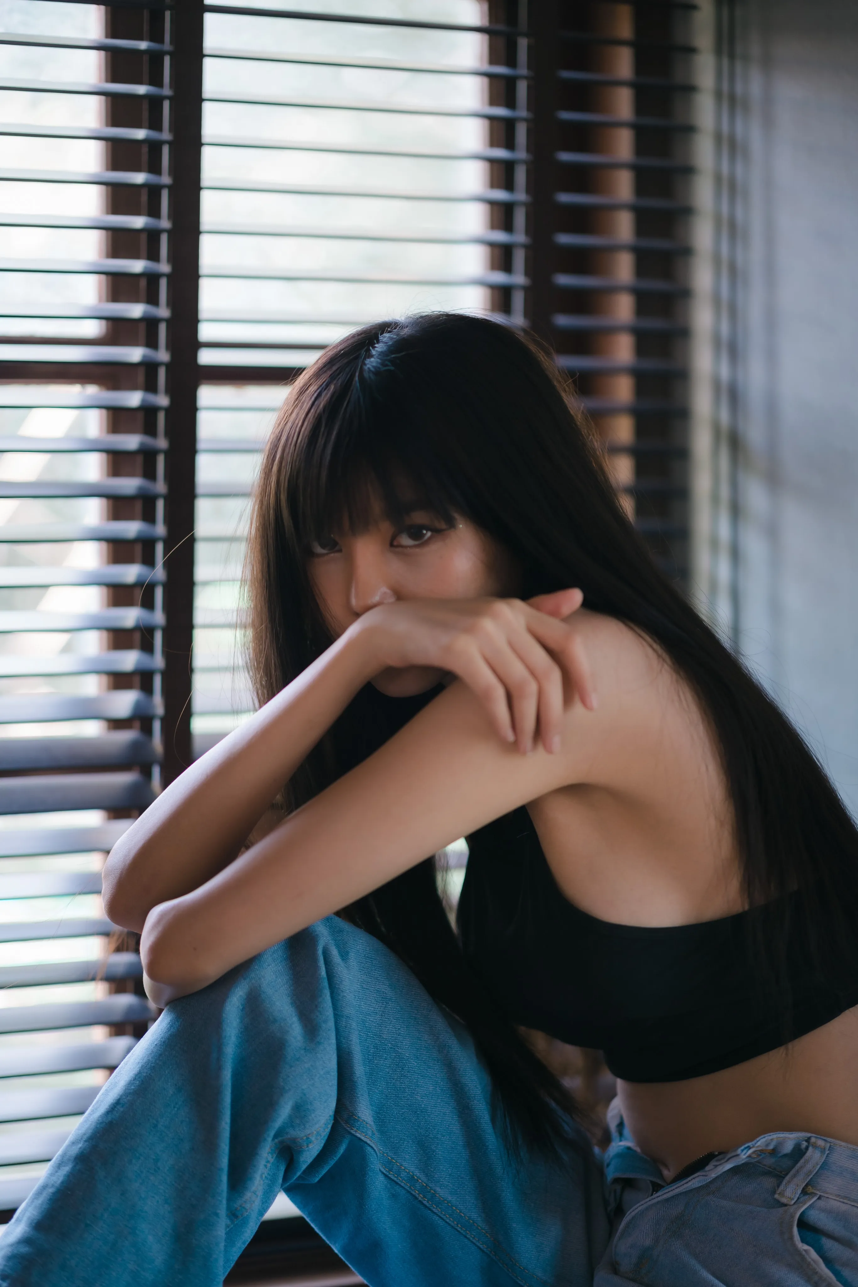 A woman with long dark hair and bangs resting her face on her folded arms beside window blinds