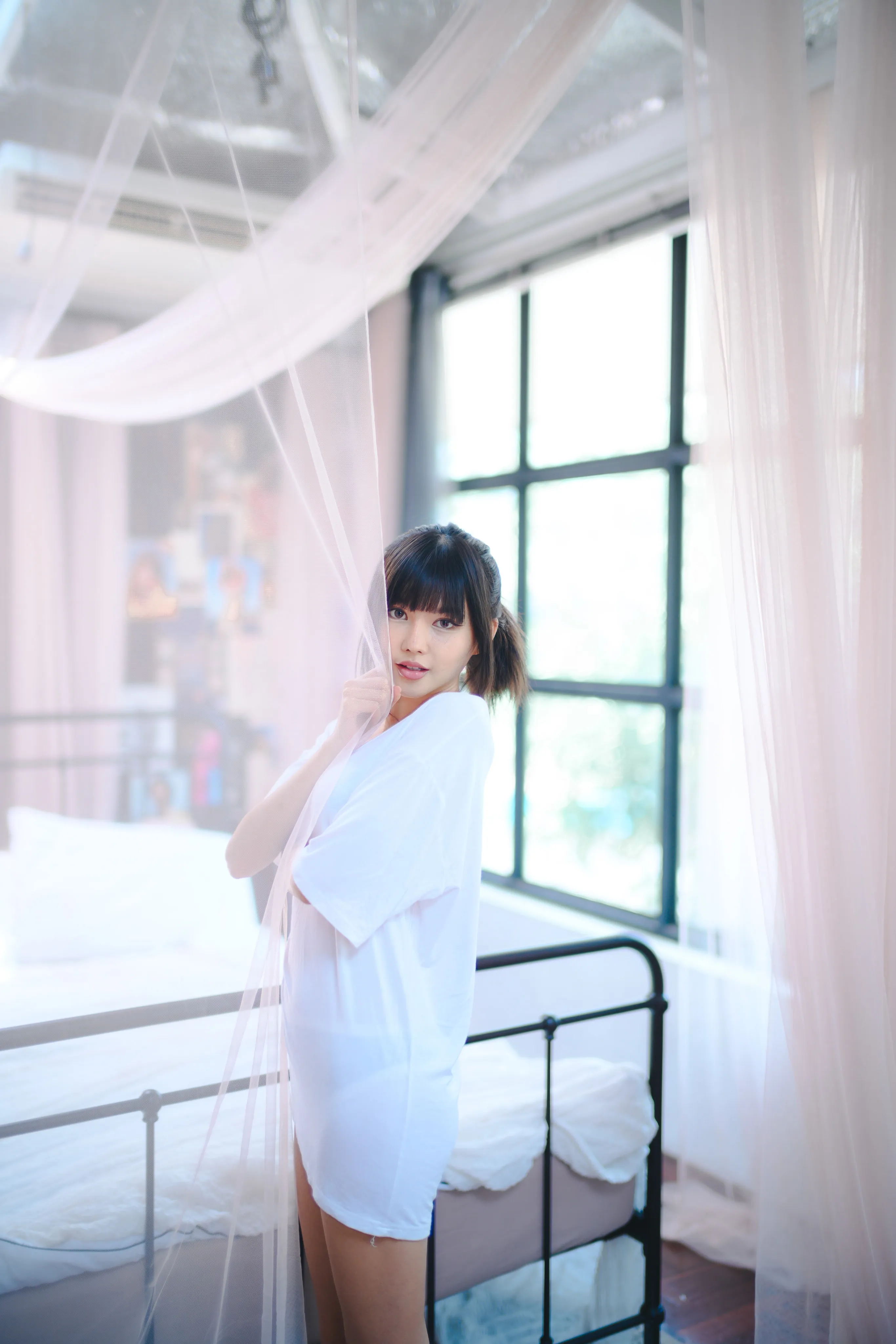 A woman in a white T-shirt standing among sheer white canopy curtains in a bright bedroom