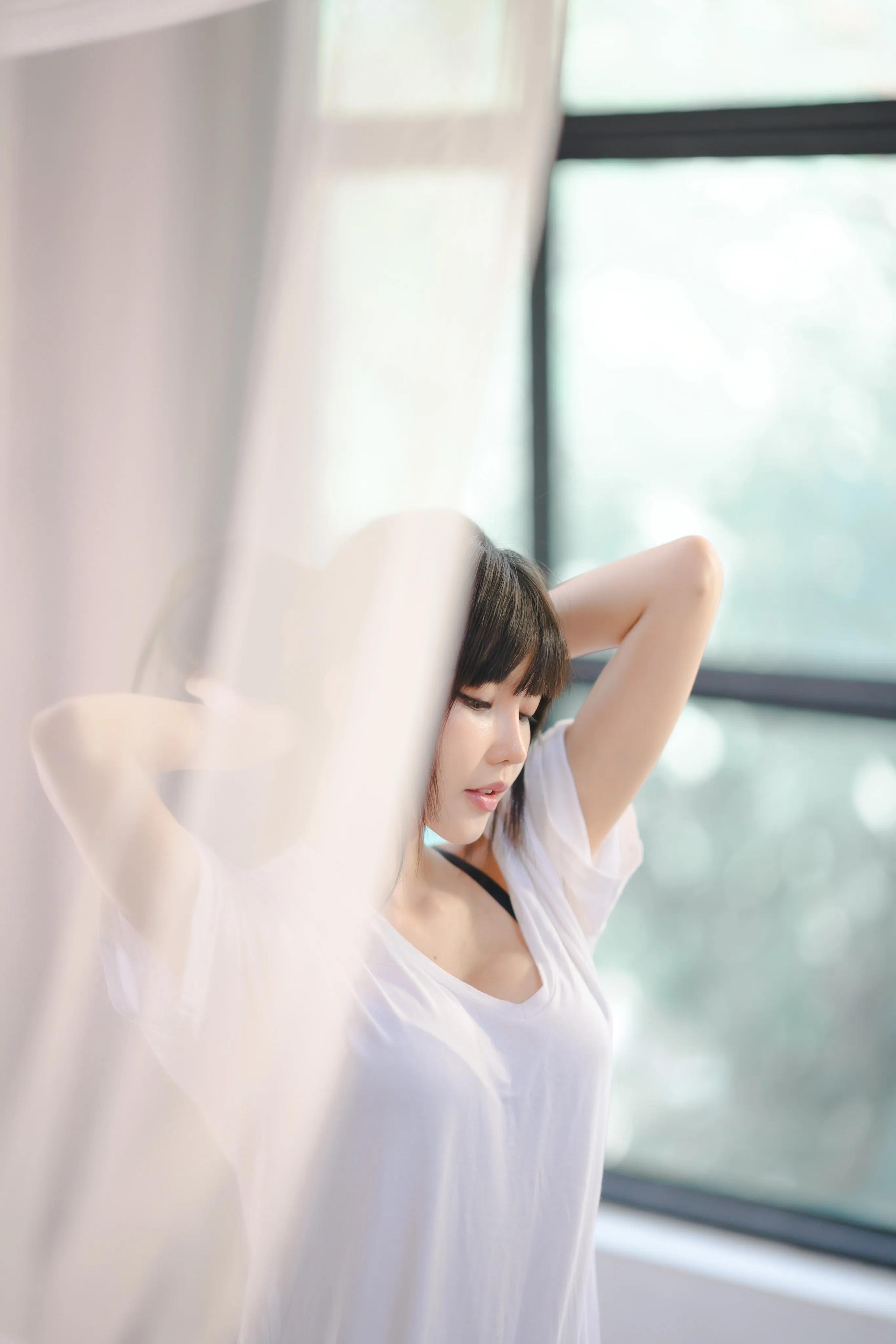 A woman in a white T-shirt raising her arms behind her head, softly framed by a sheer curtain