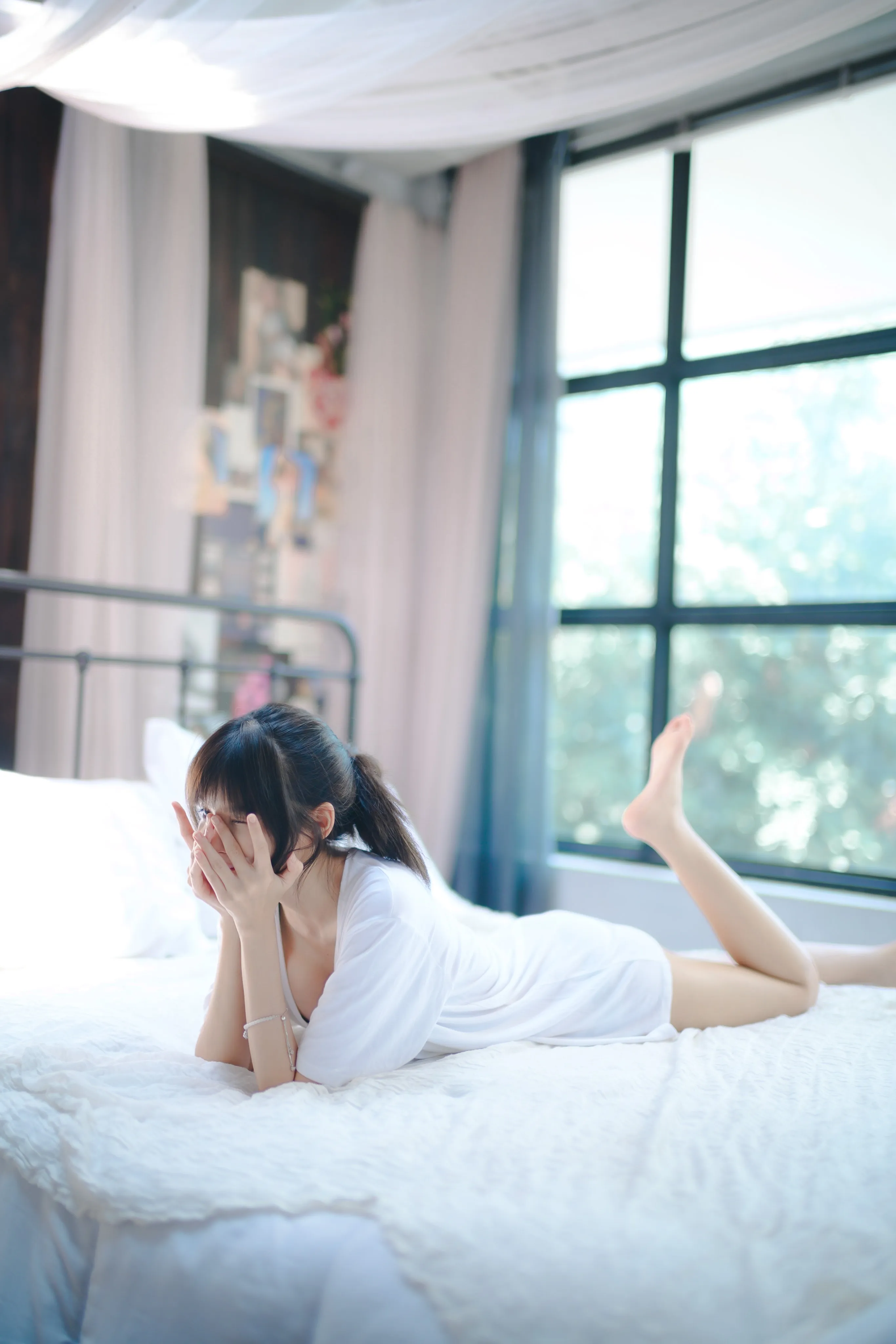 A woman in a white T-shirt lying on a bed covering her face with her hands, feet raised