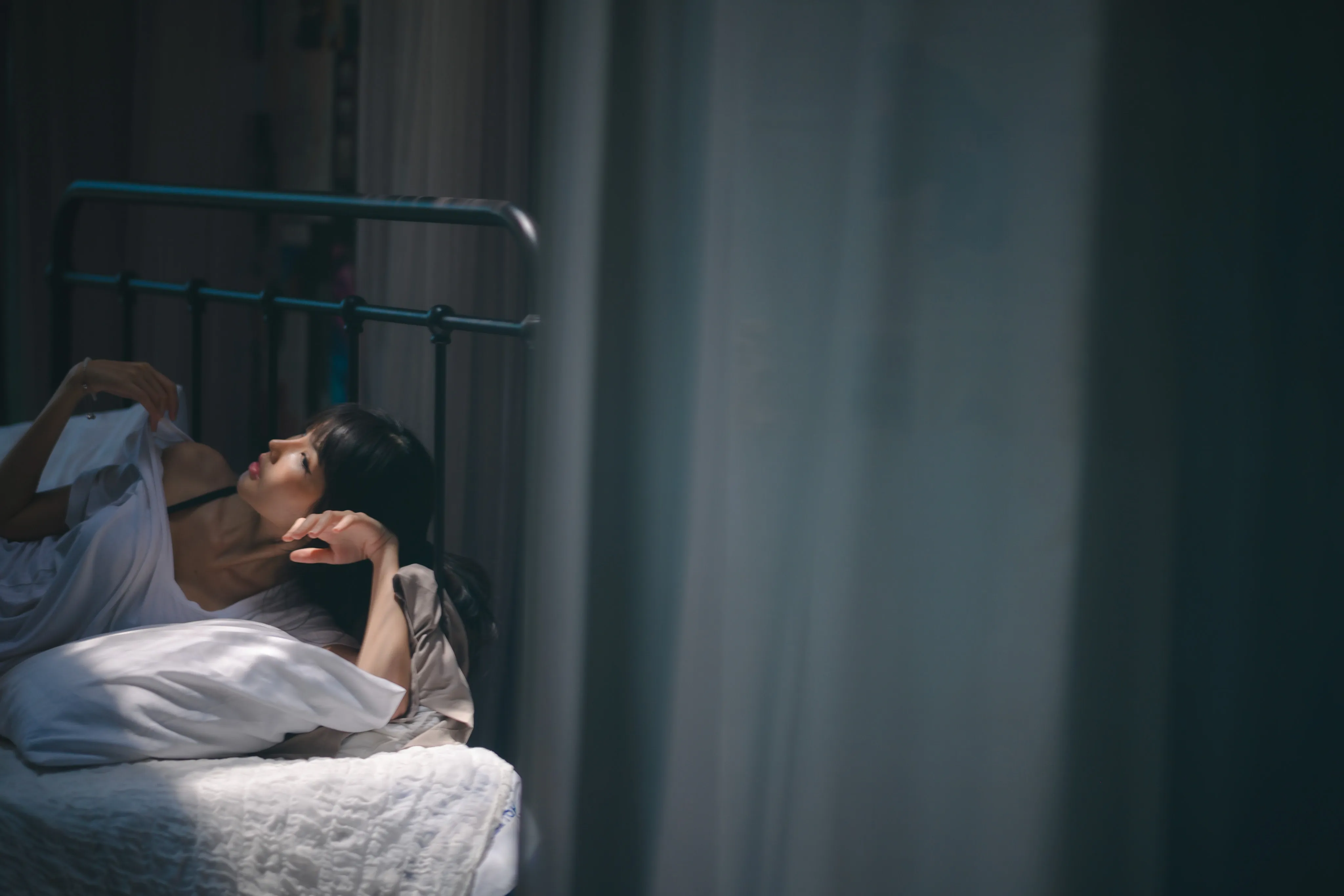 A woman lying on a pillow on an iron-framed bed in a dim, blue-toned room