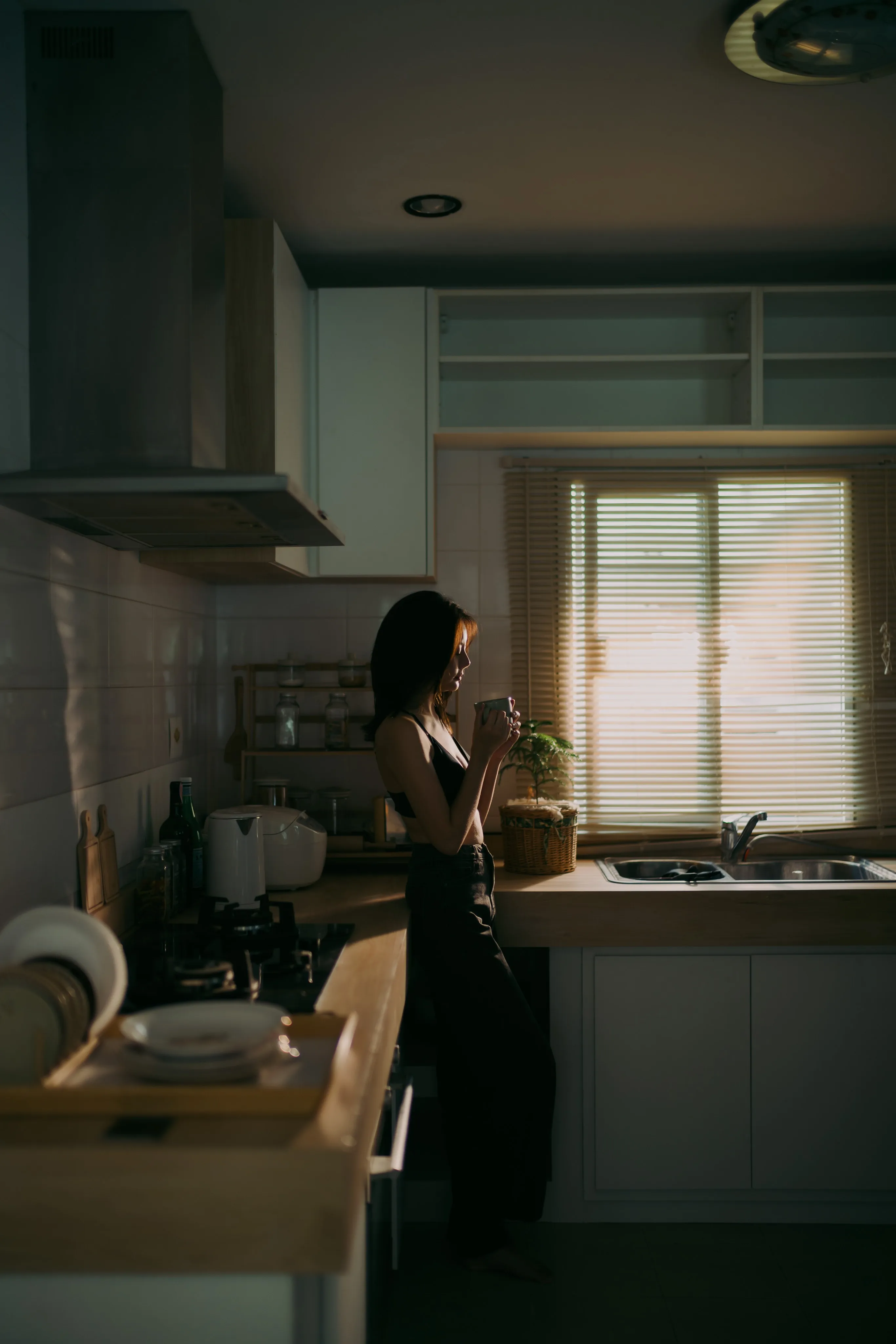 A woman holding a mug in a dim kitchen in front of a window with blinds