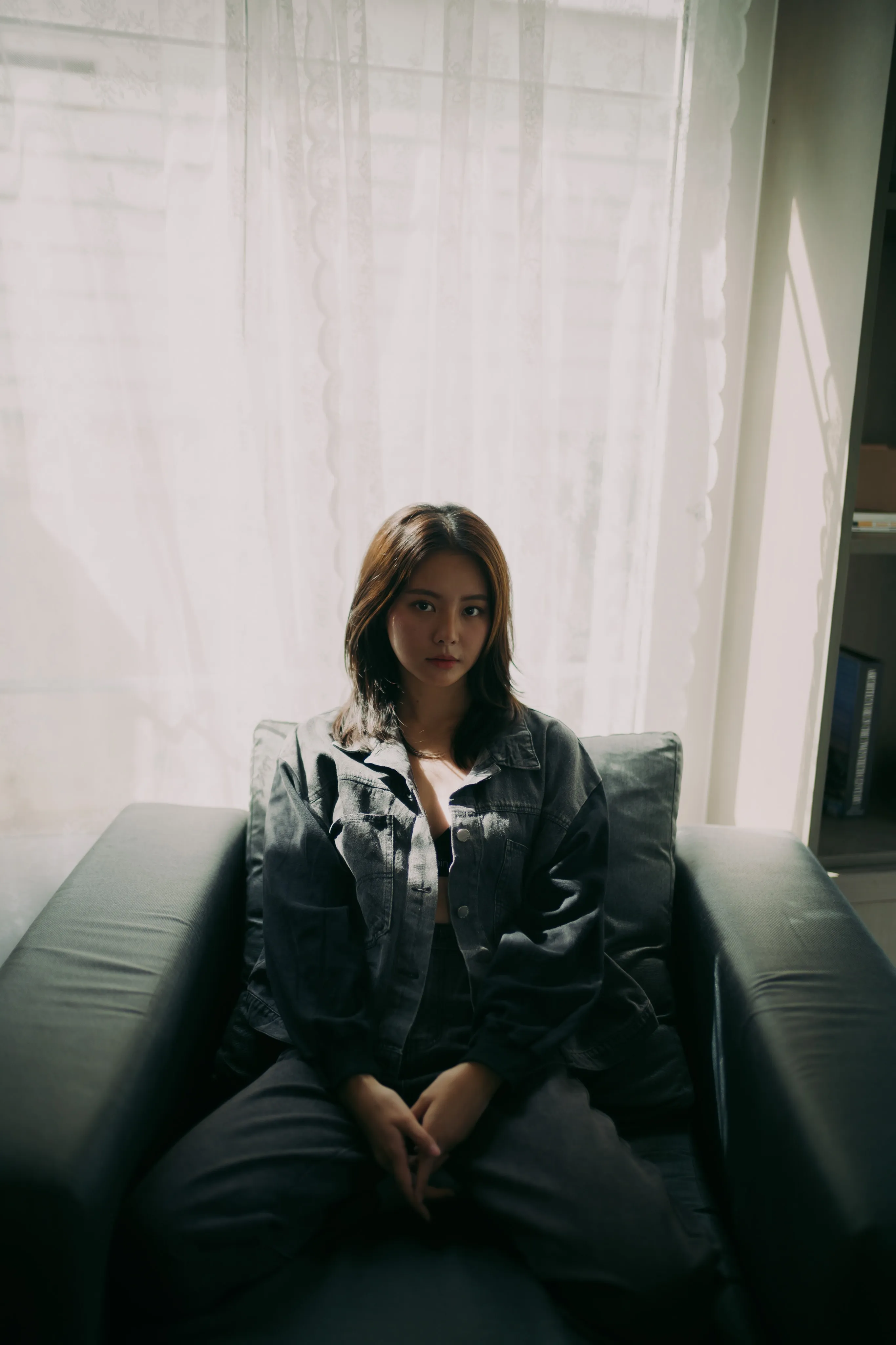 A woman in a denim jacket sitting in a dark armchair in front of sheer white curtains