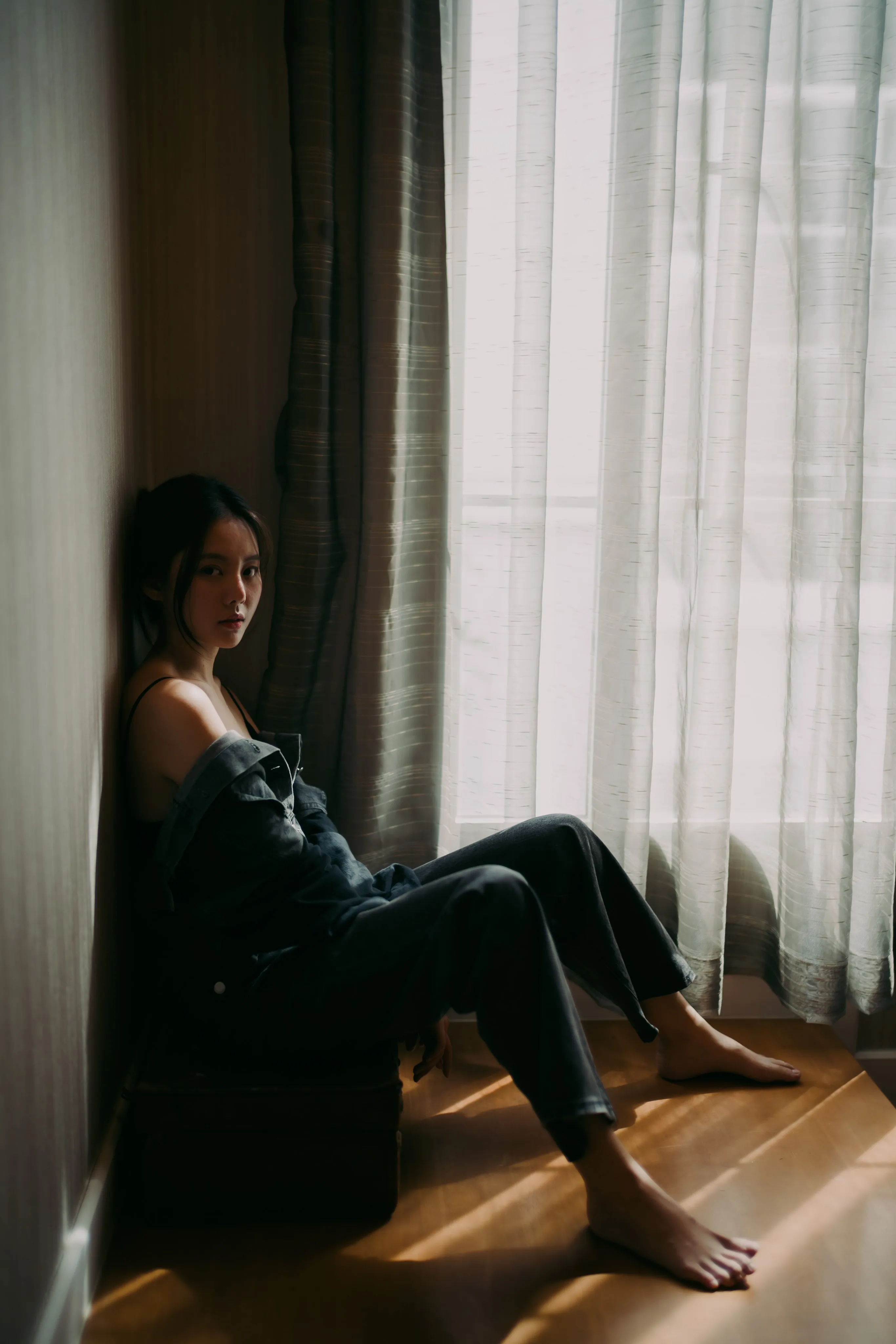 A woman in denim sitting on the floor against a wall beside curtains, in warm window light