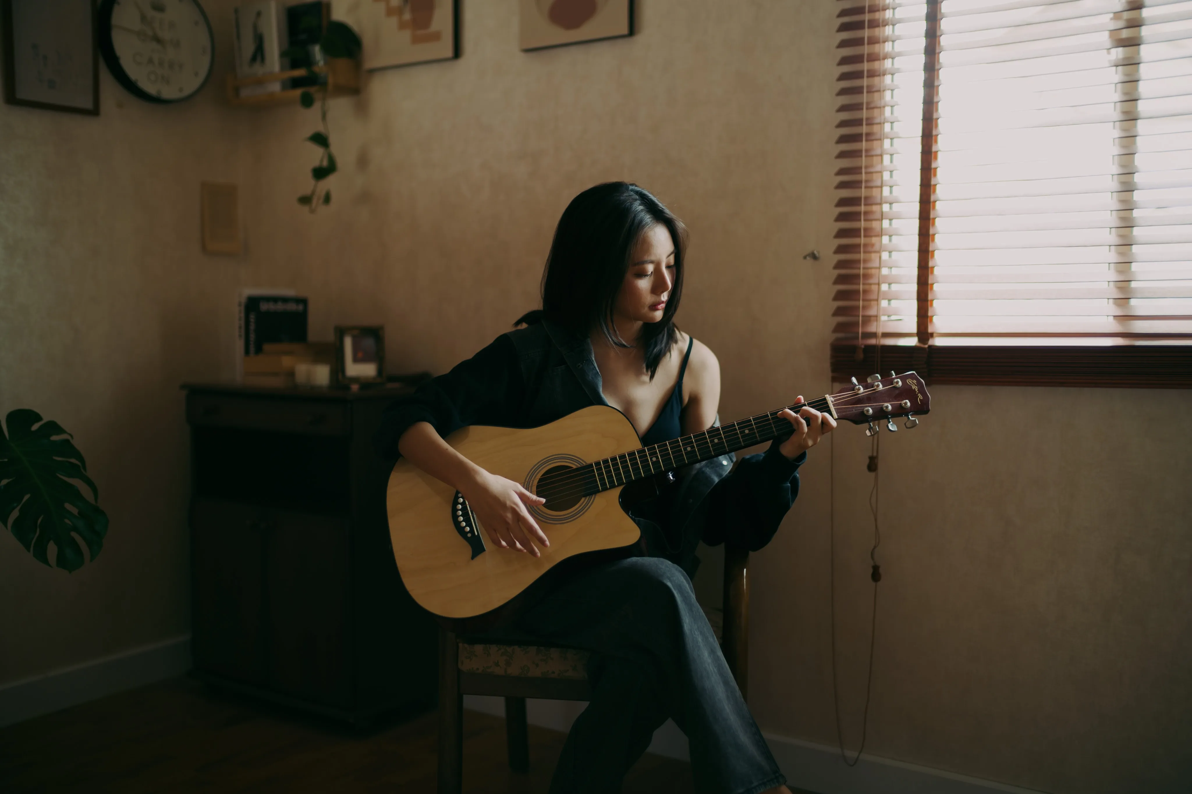 A woman in dark clothing playing an acoustic guitar beside a window with wooden blinds