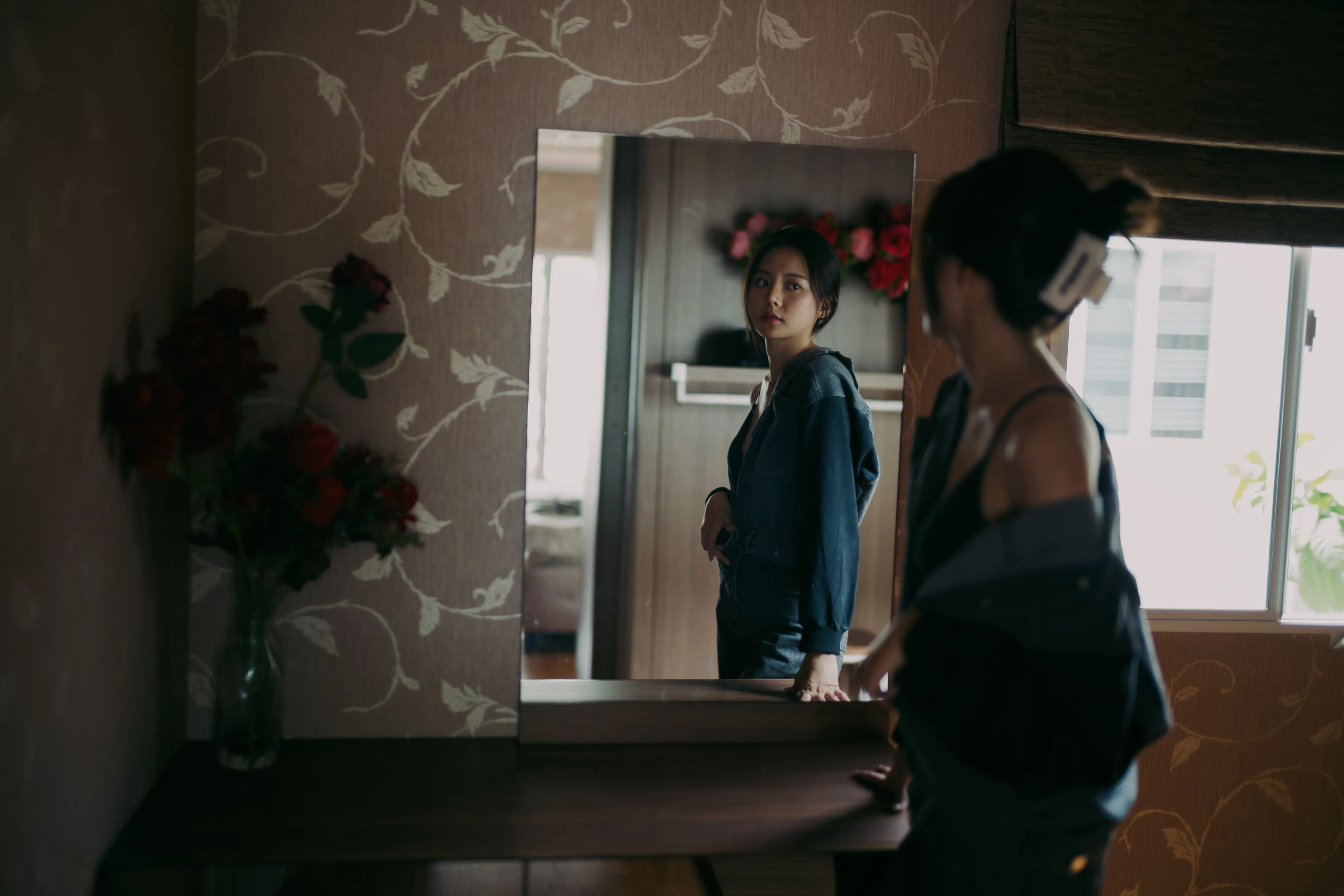 A woman in denim glancing at her reflection in a mirror in a room with patterned wallpaper