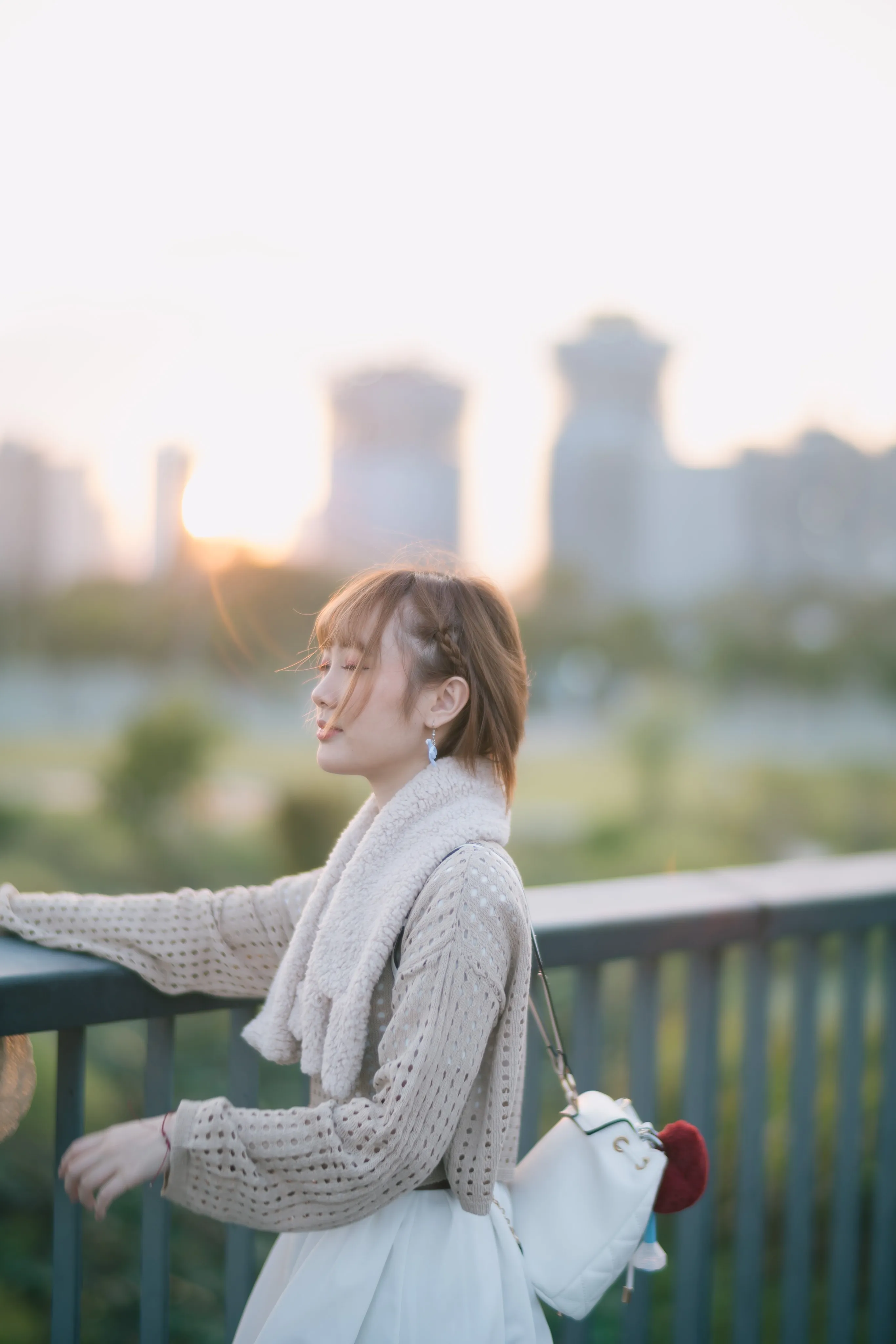 A young woman in a knit cardigan leaning on a railing with a blurred city skyline at sunset
