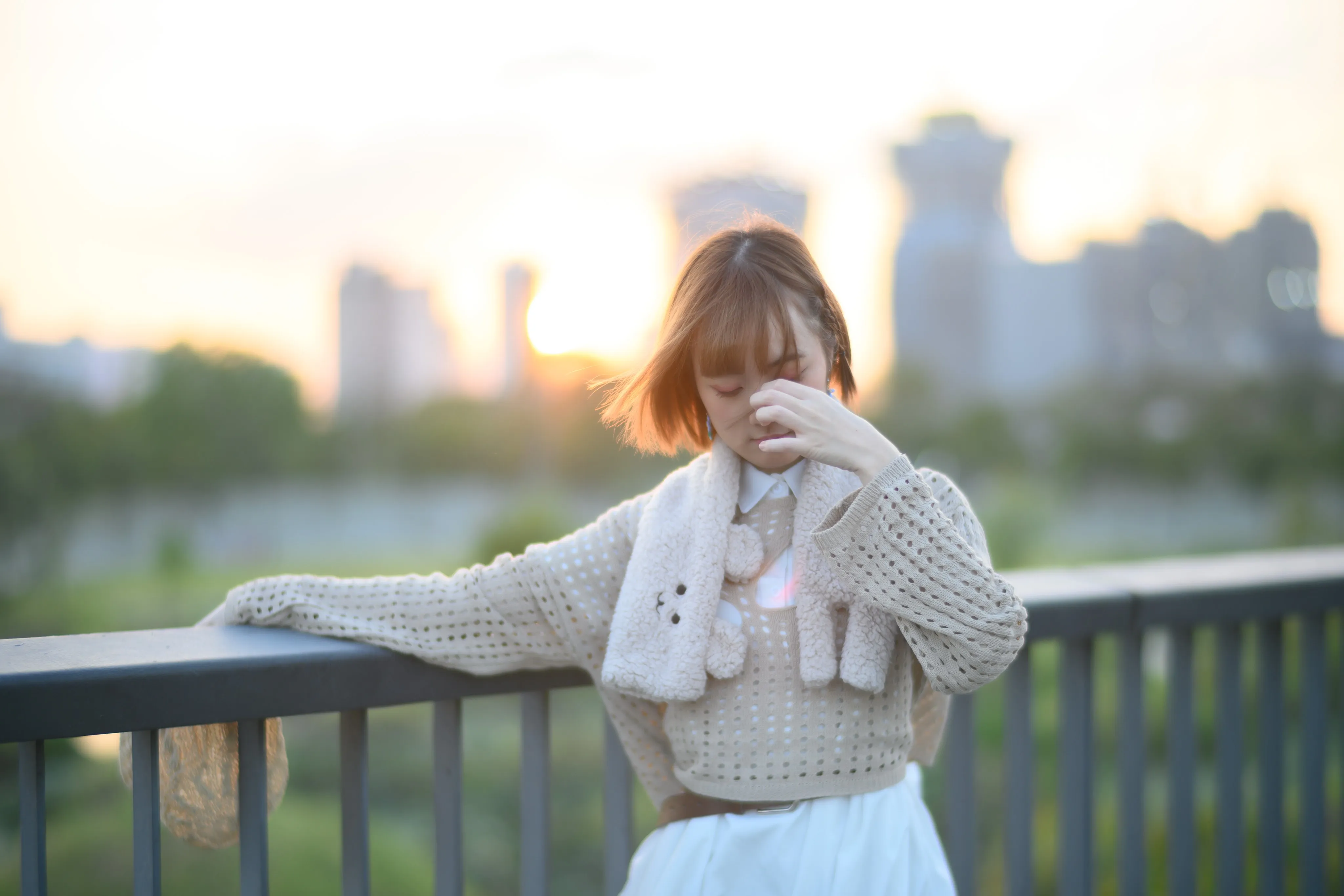 A young woman in a knit cardigan touching her face at a railing, backlit by the setting sun over the city