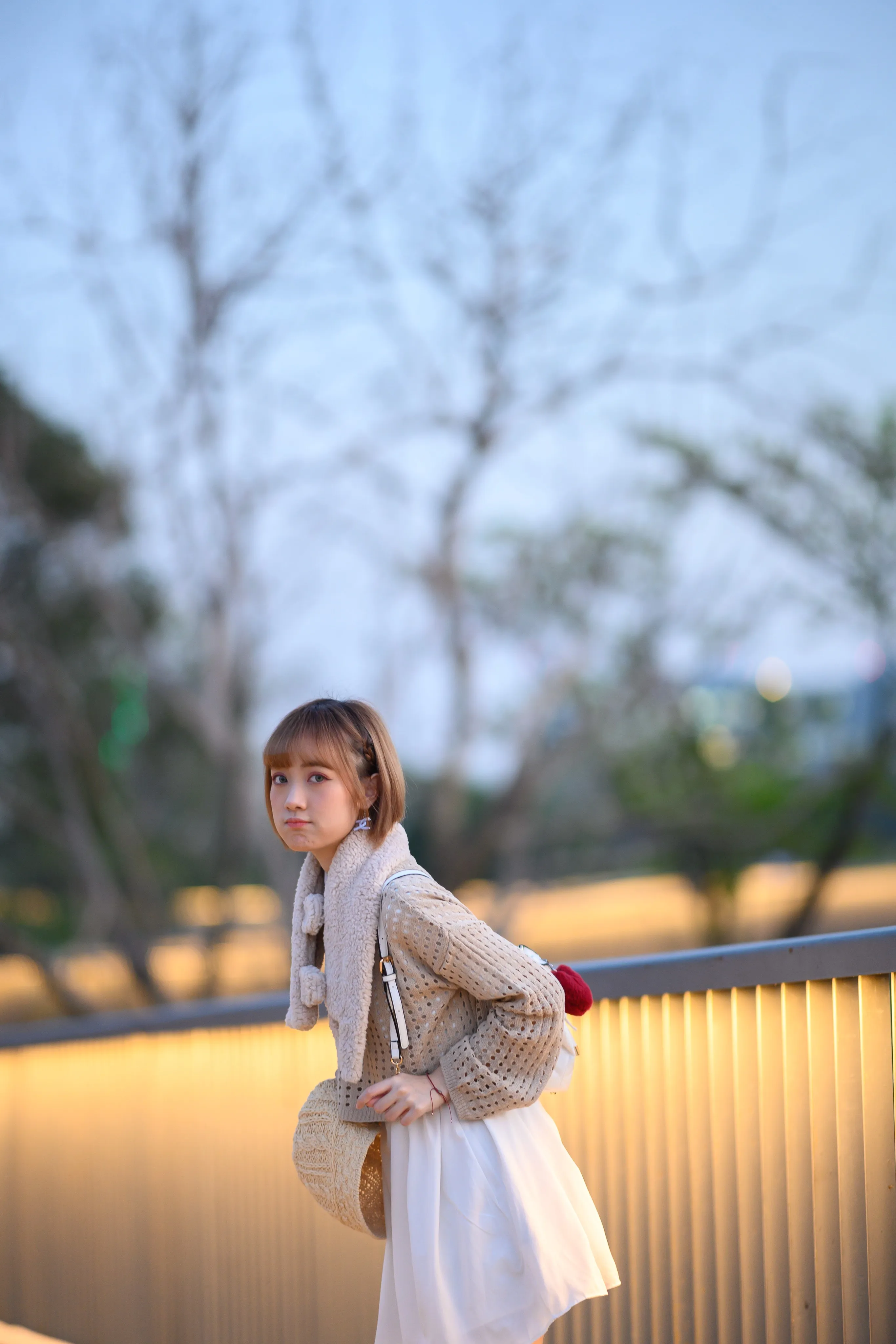A young woman in a knit cardigan and white skirt looking back over her shoulder at a lit railing at dusk