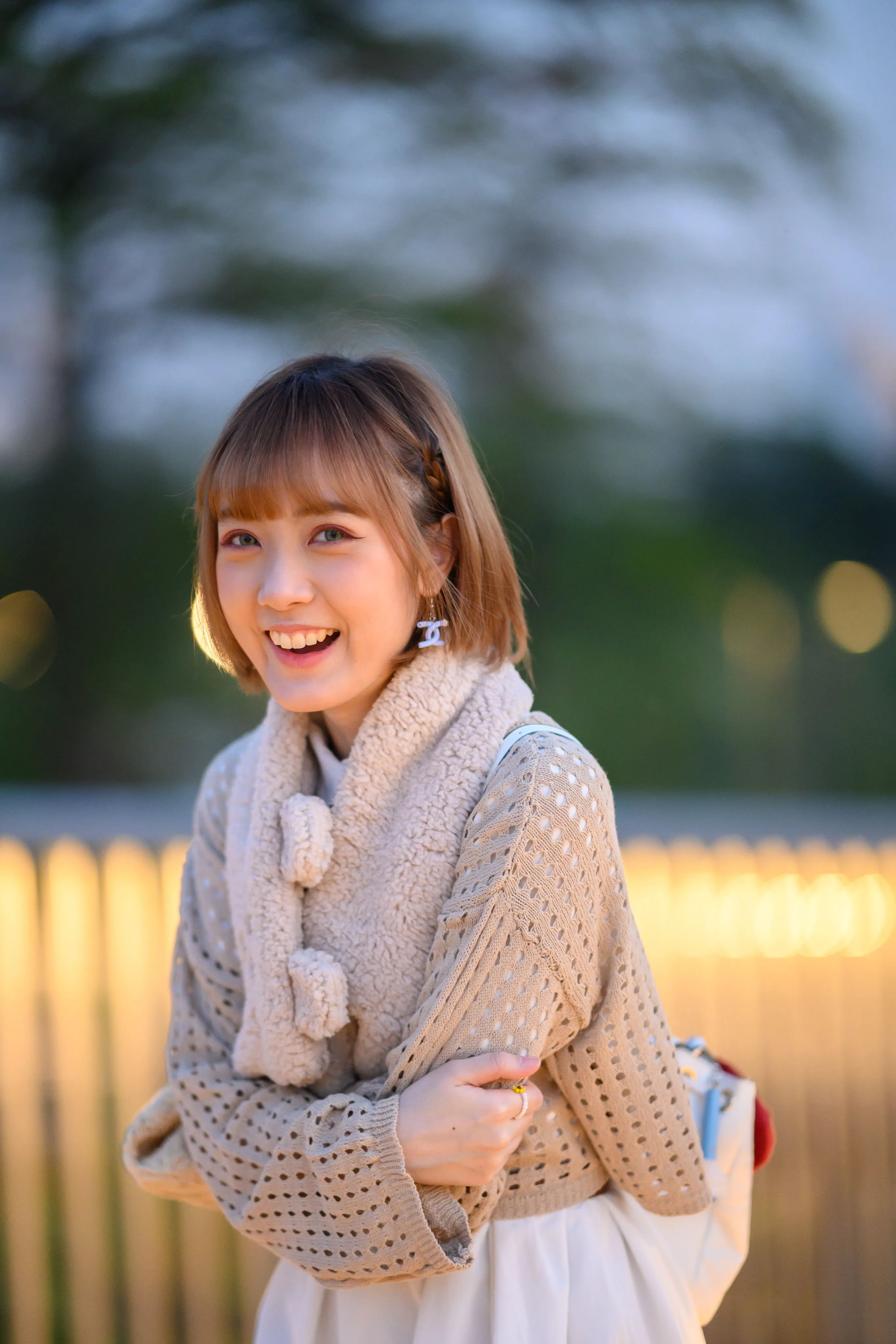 A young woman in a knit cardigan and pom-pom scarf laughing in front of warm evening lights