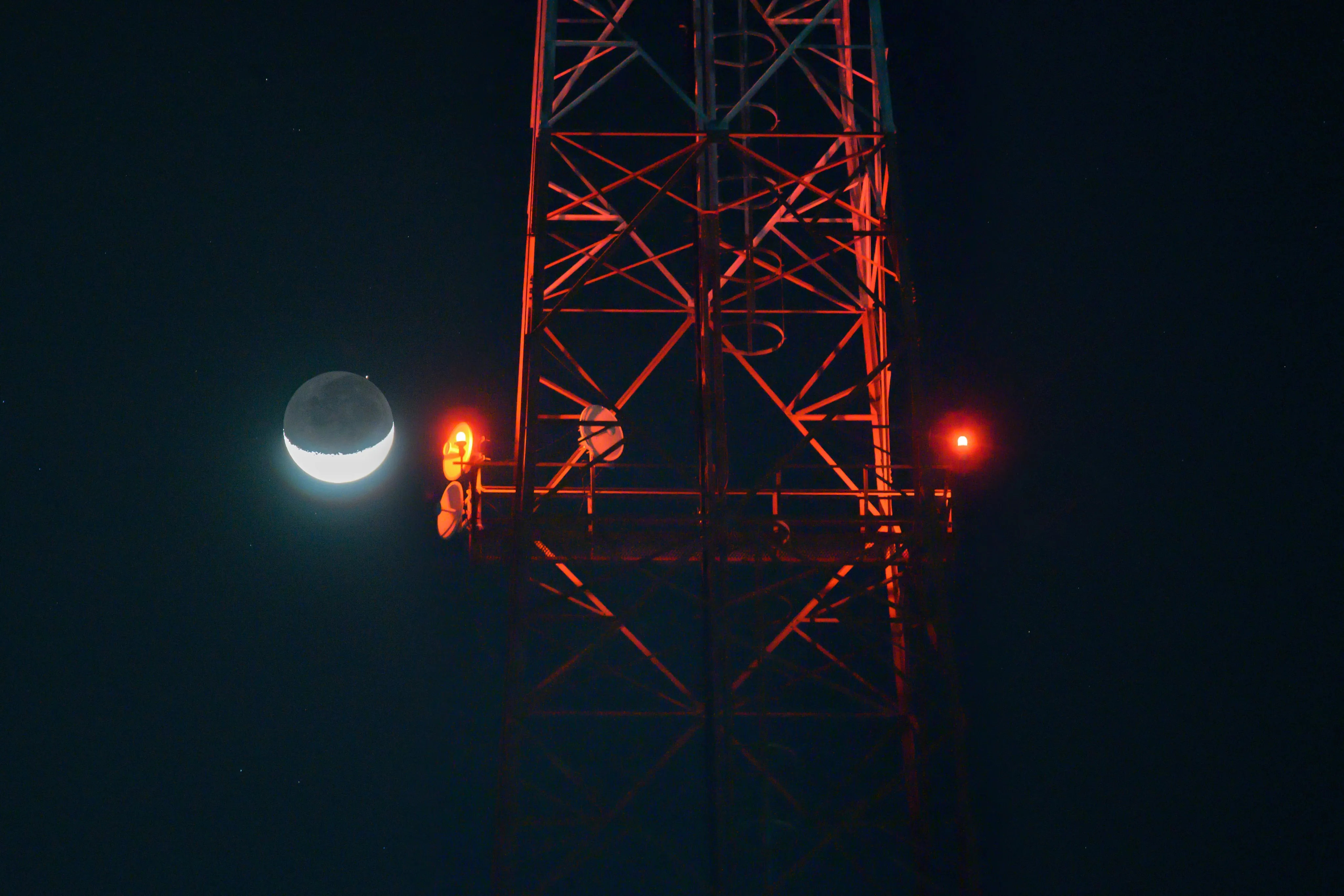 A thin crescent moon beside a red-lit steel transmission tower at night