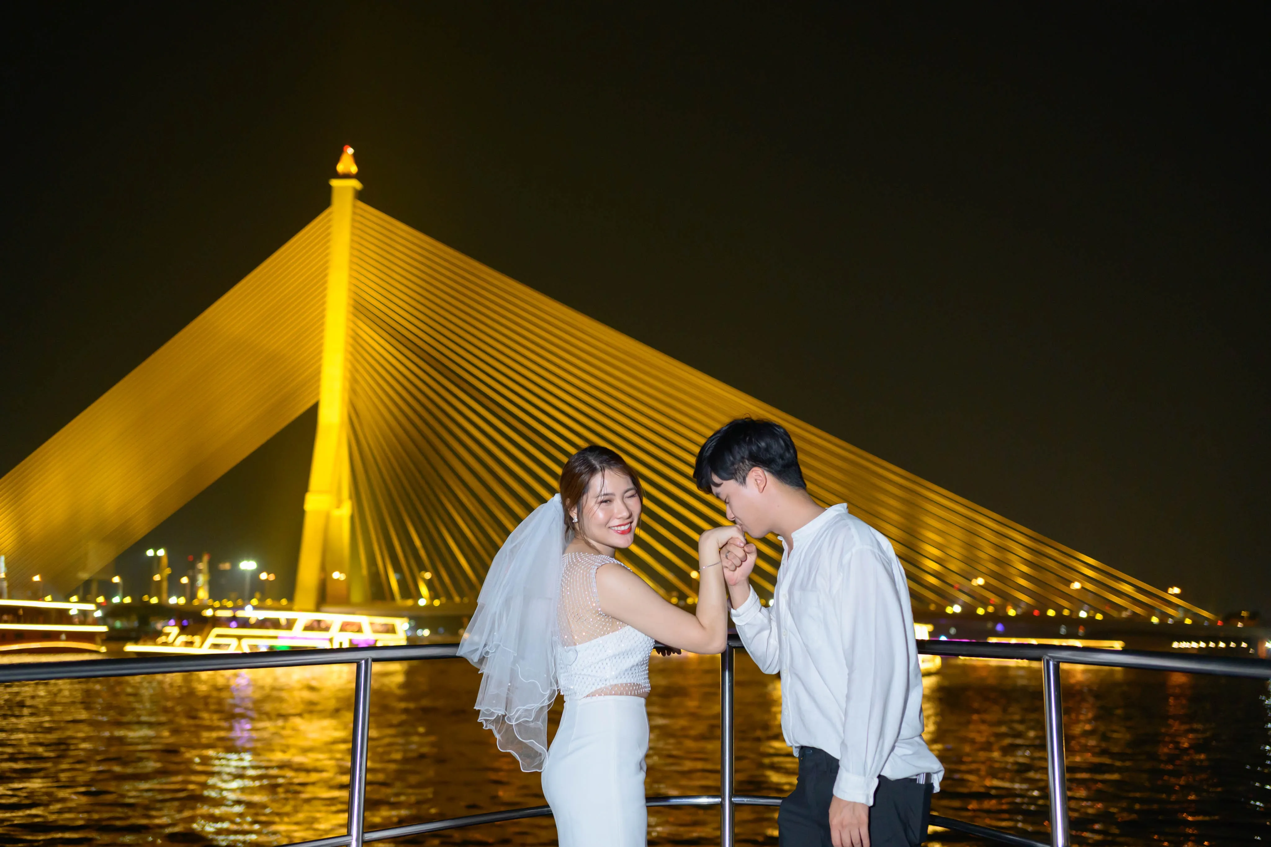 A couple holding hands on a boat deck in front of a golden-lit cable-stayed bridge at night