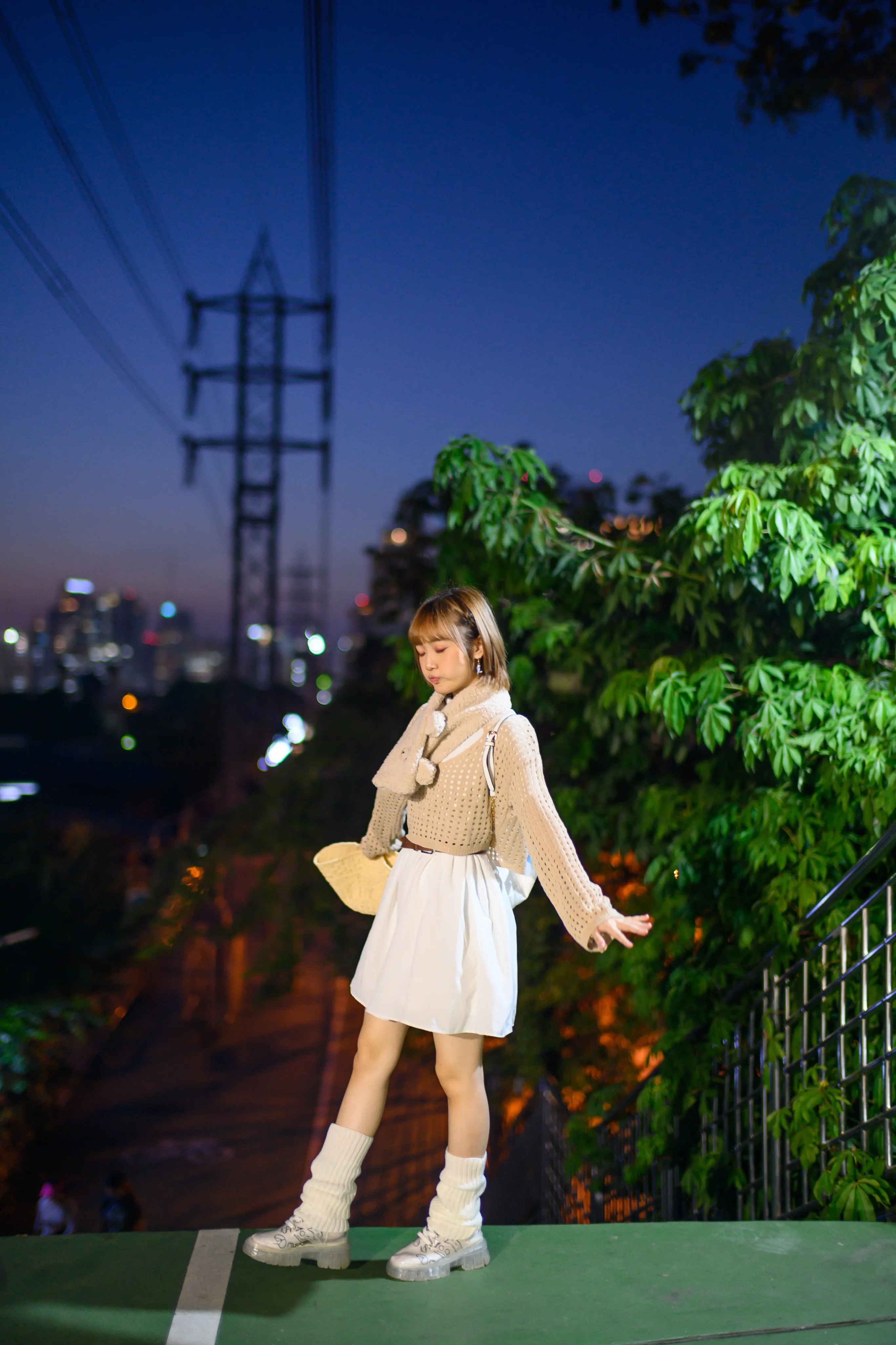 A young woman in a knit cardigan and white skirt twirling on a path under power lines at dusk