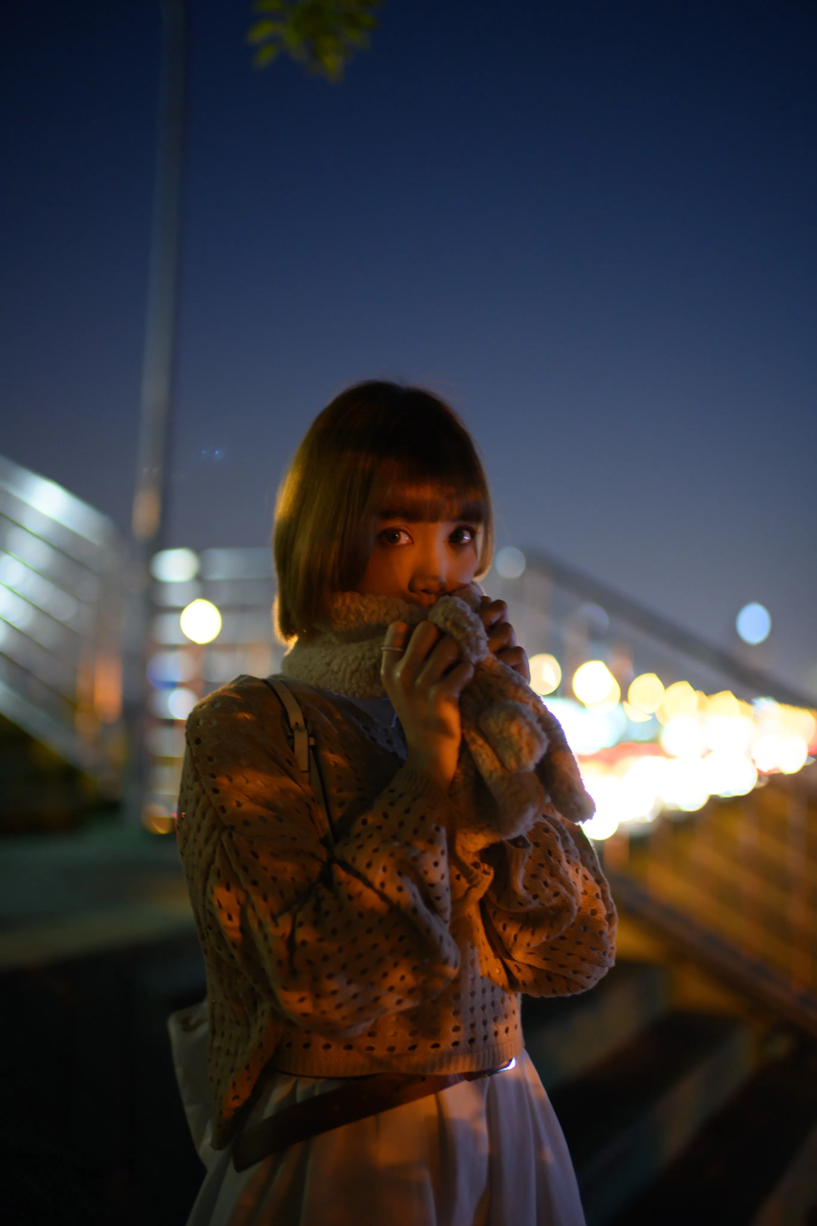 A young woman holding her scarf up to her face on a footbridge at night