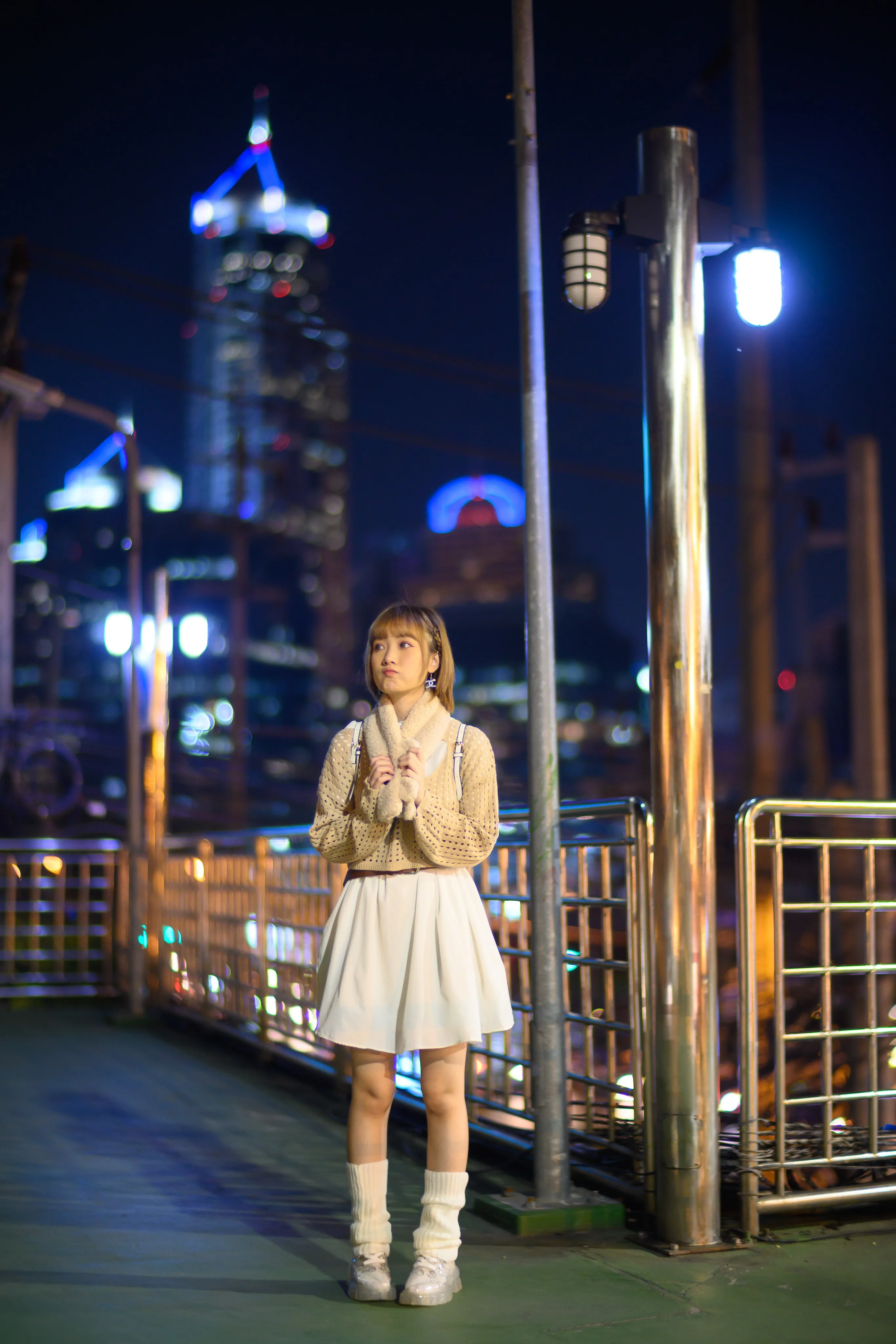 A young woman in a knit cardigan and white skirt standing on a footbridge with city towers lit at night