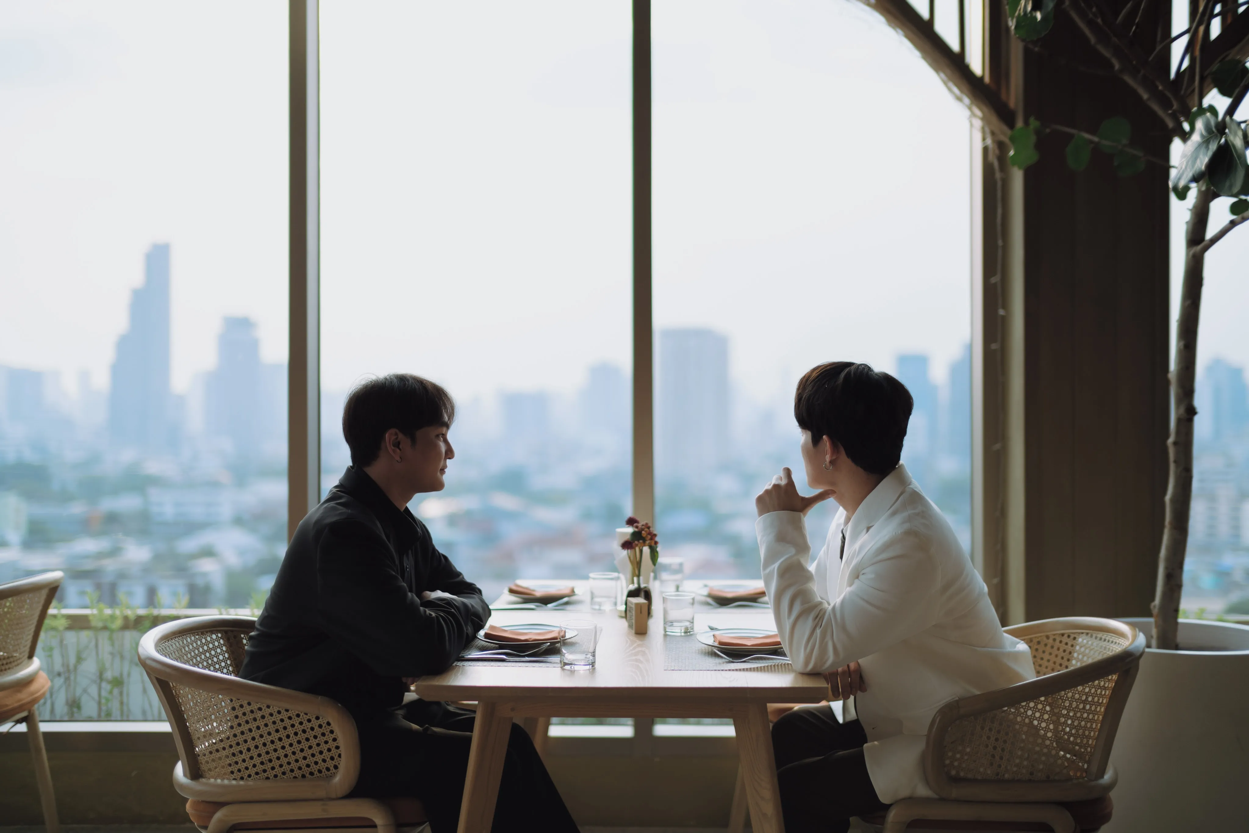 Two men in black and white jackets talking at a table by tall windows overlooking the city