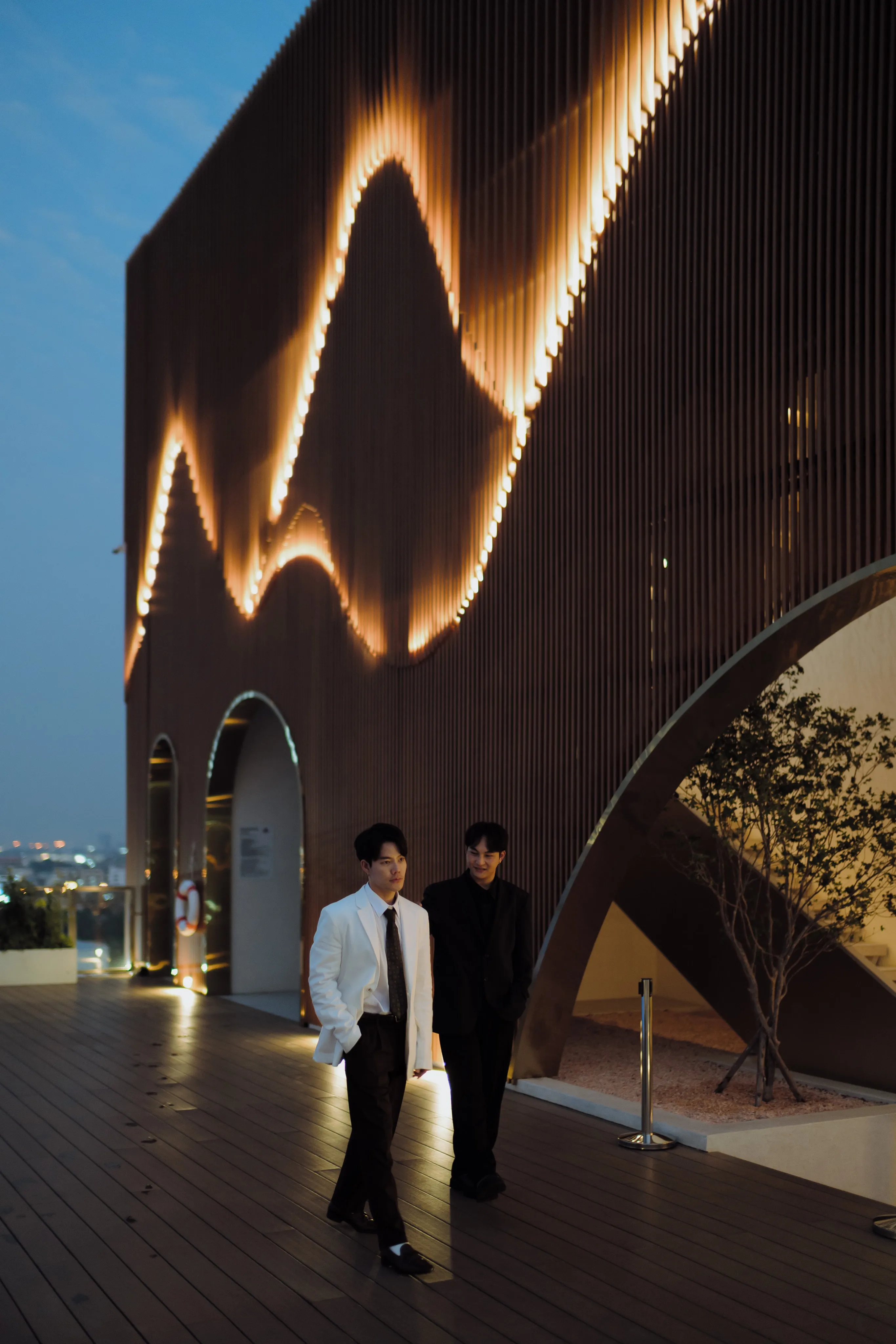Two men in white and black jackets walking along a rooftop deck beside a wall of glowing lights