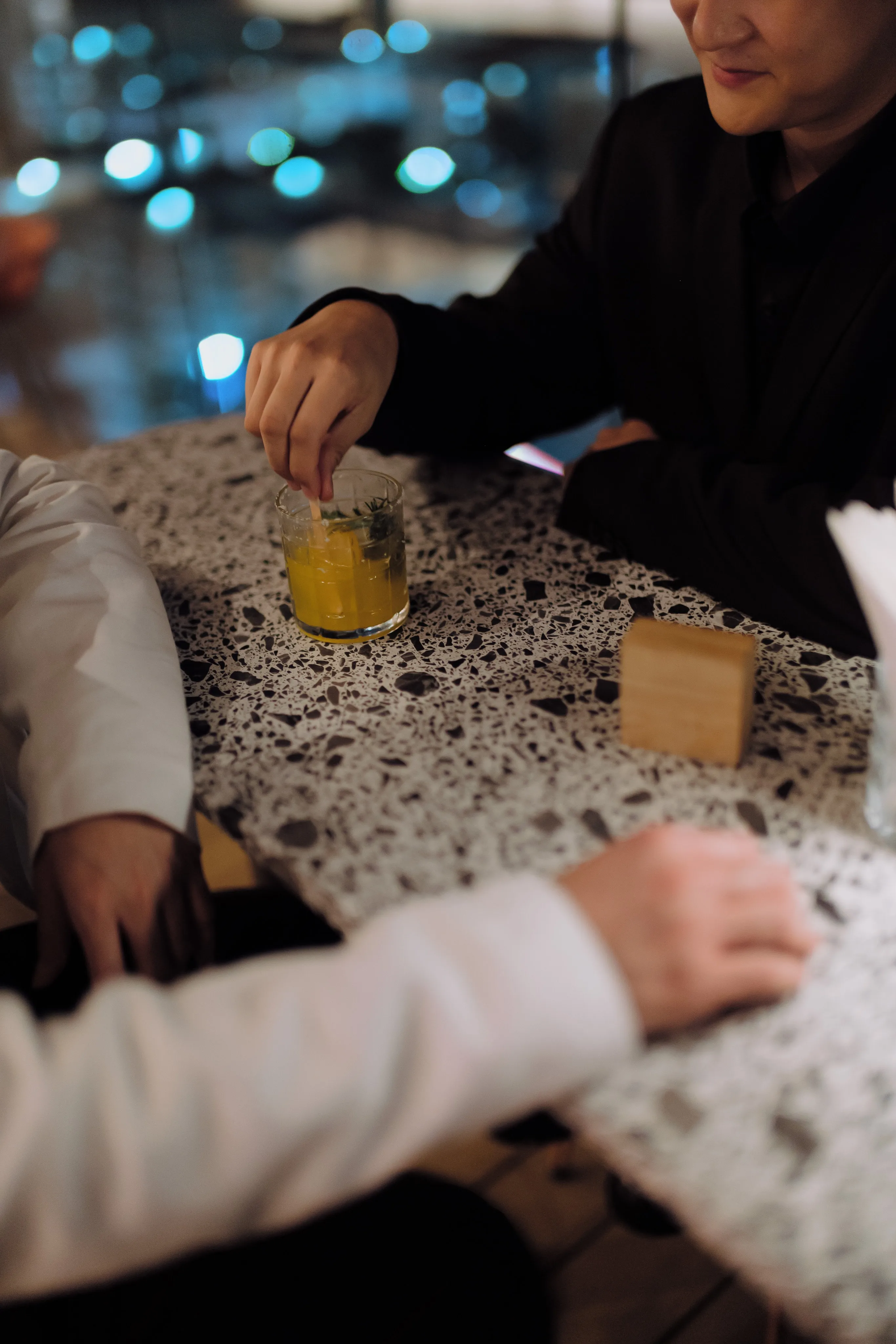 Close-up of hands at a terrazzo table, one stirring a yellow drink